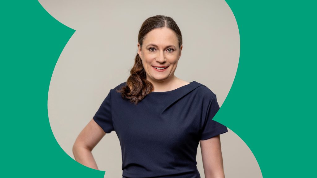 Portrait of Dr. Daniela Büchel. She has one arm on her hip and is smiling at the camera. The background is gray with green details.
