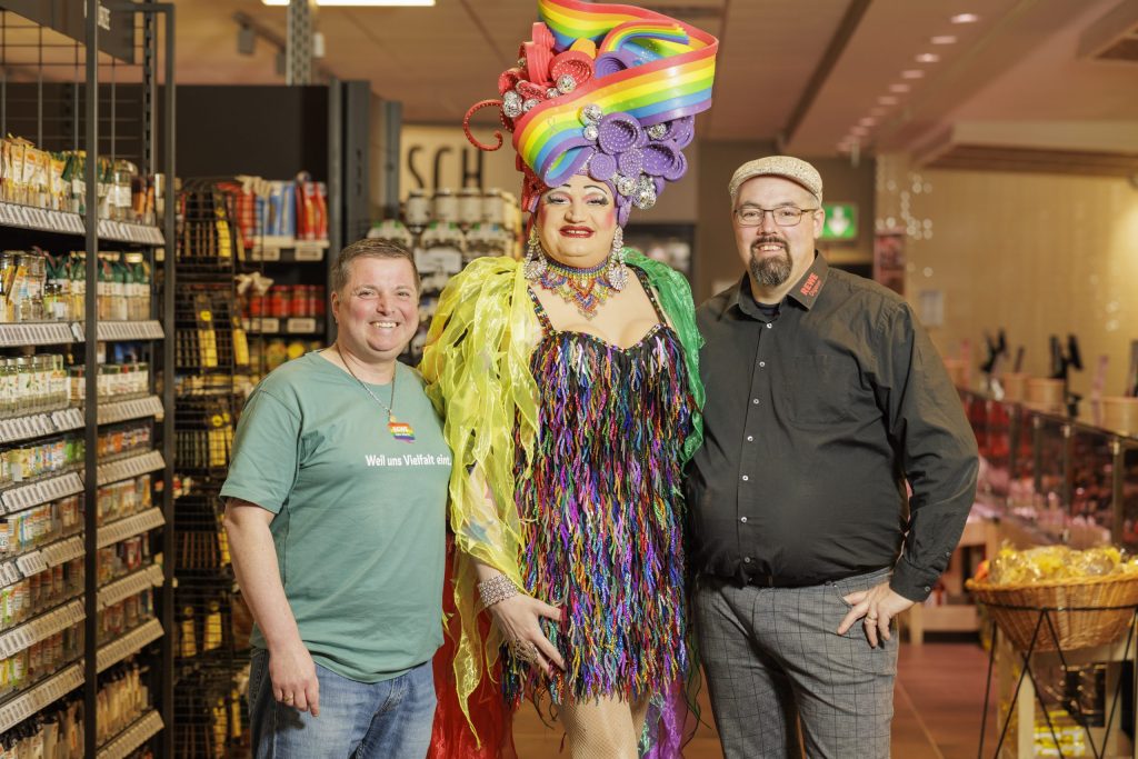 Group photo of REWE salesman Jérôme Digeon with a colleague and a member of a travesty group styled as a drag queen. The group is standing in a REWE store.