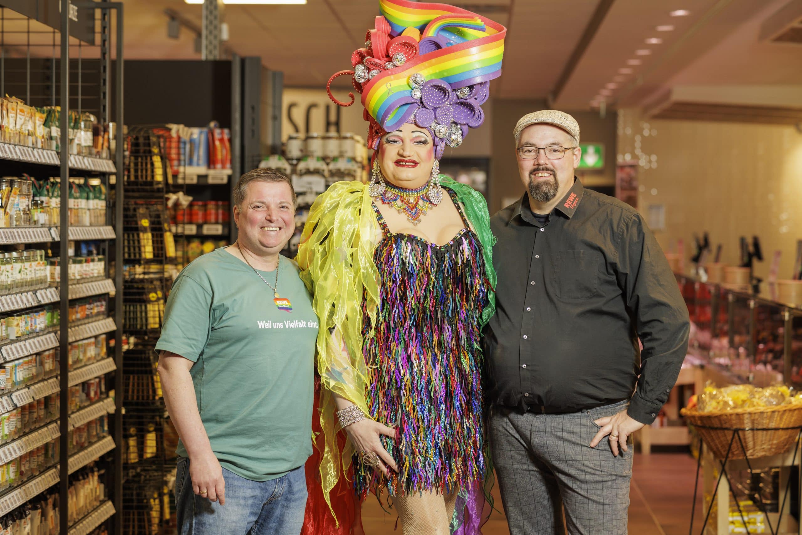 Gruppenbild von REWE Kaufmann Jérôme Digeon mit einem Kollegen und einem Mitglied aus einer Travestiegruppe als Dragqueen gestyled. Die Gruppe steht in einem REWE Markt.