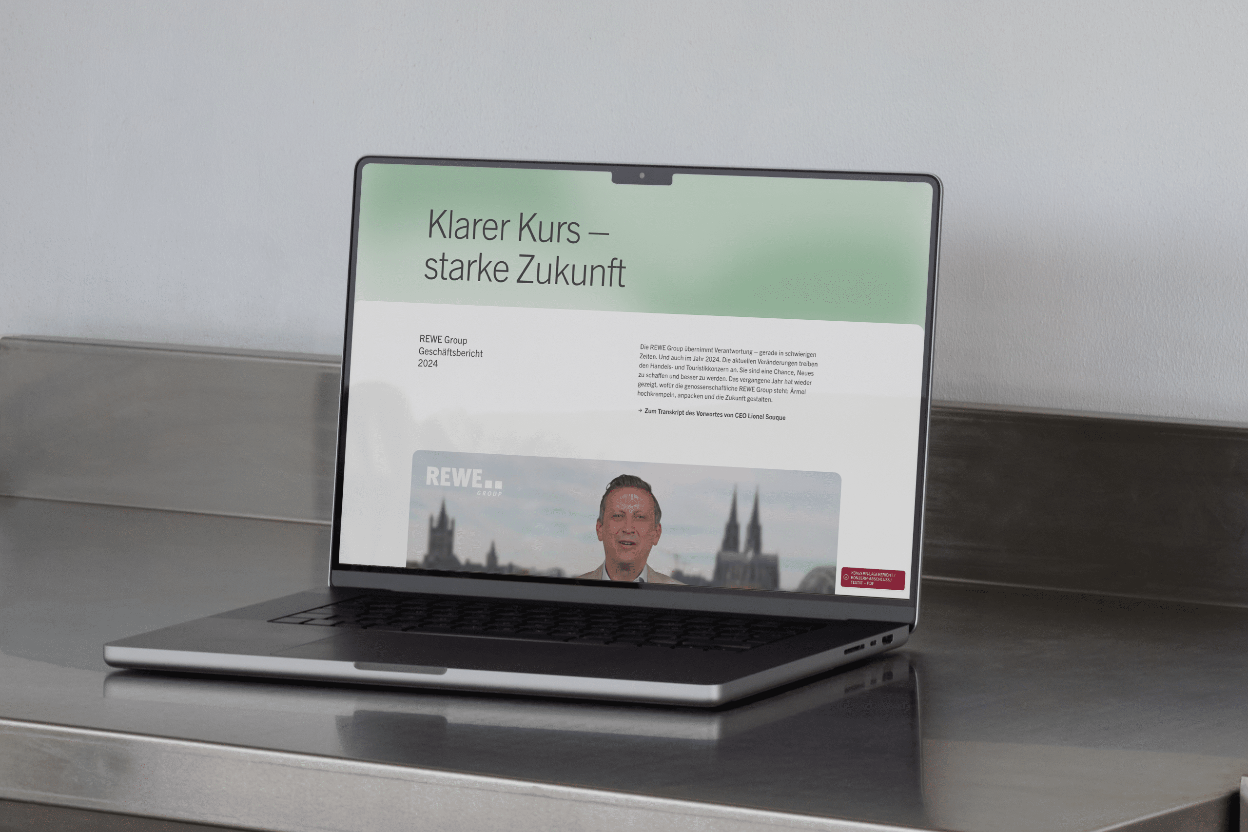 Geöffneter Laptop auf einer Ablage aus Edelstahl. Auf dem Bildschirm ist die Startseite des Geschäftsberichts der REWE Group 2024.