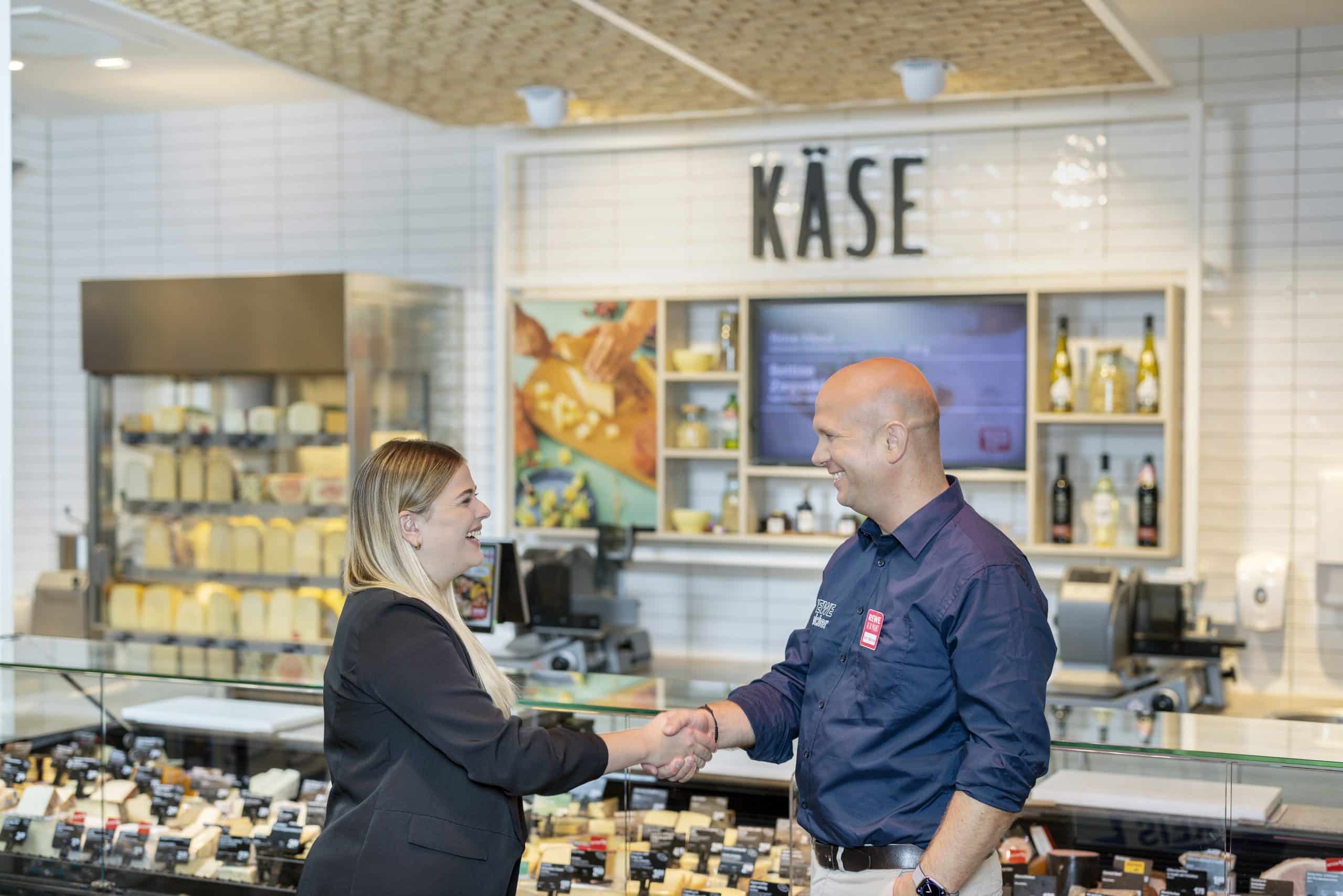 Two people are standing in front of the cheese counter in a REWE supermarket, laughing and shaking hands.