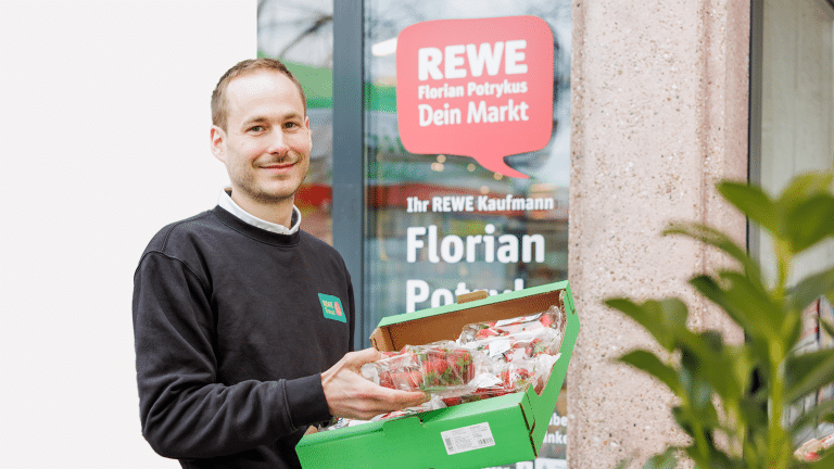 Porträt von Konstantin Tönnies. Er hält einen Karton mit Erdbeeren in der Hand und lächelt in die Kamera. Im Hintergrund ist der Eingang eines REWE Marktes zu sehen.