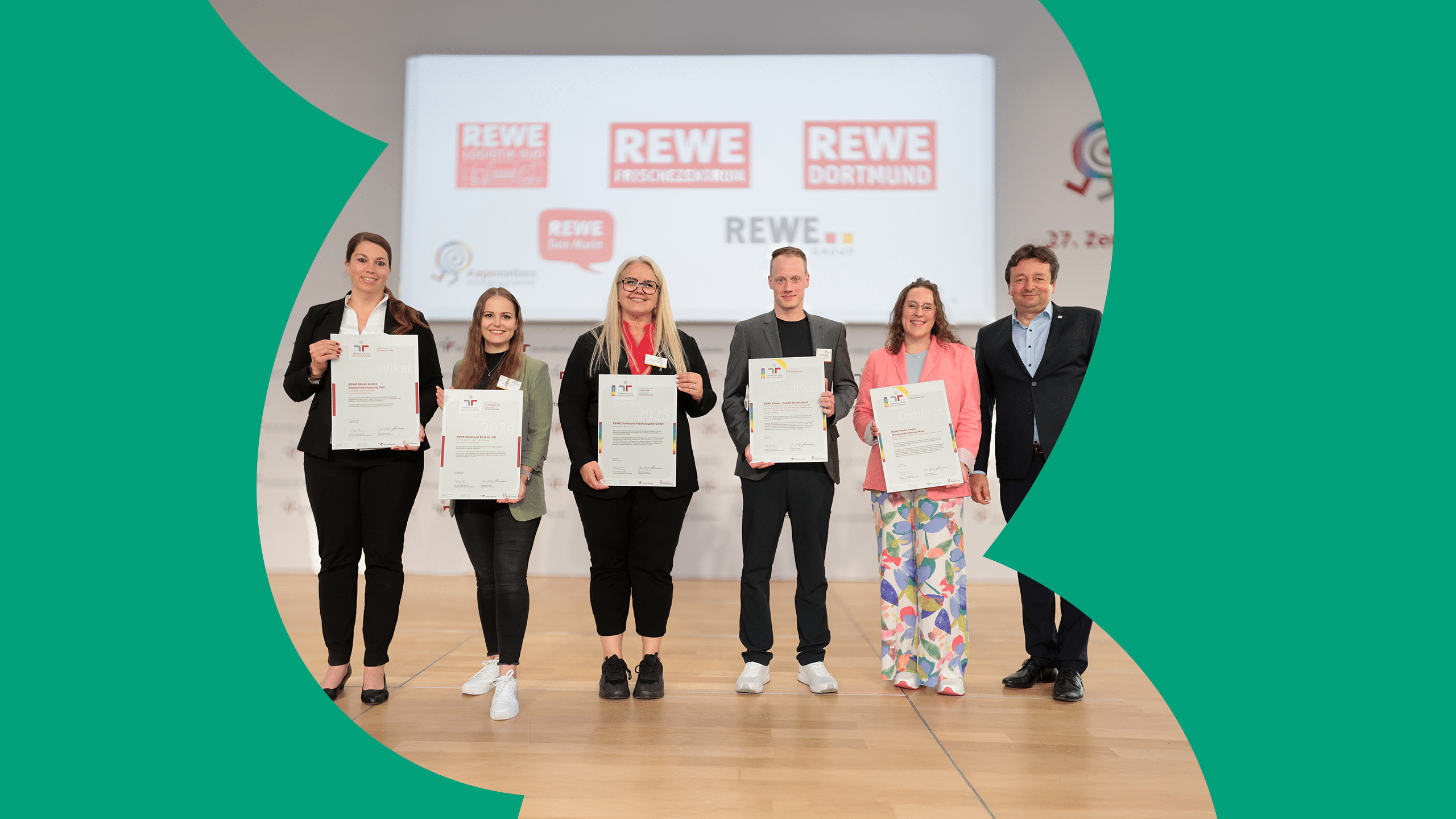 Sechs Personen stehen auf einer Bühne, fünf von Ihnen halten ein Zertifikat in der Hand. Im Hintergrund ist das REWE Logo der zertifizierten Bereiche zu sehen.