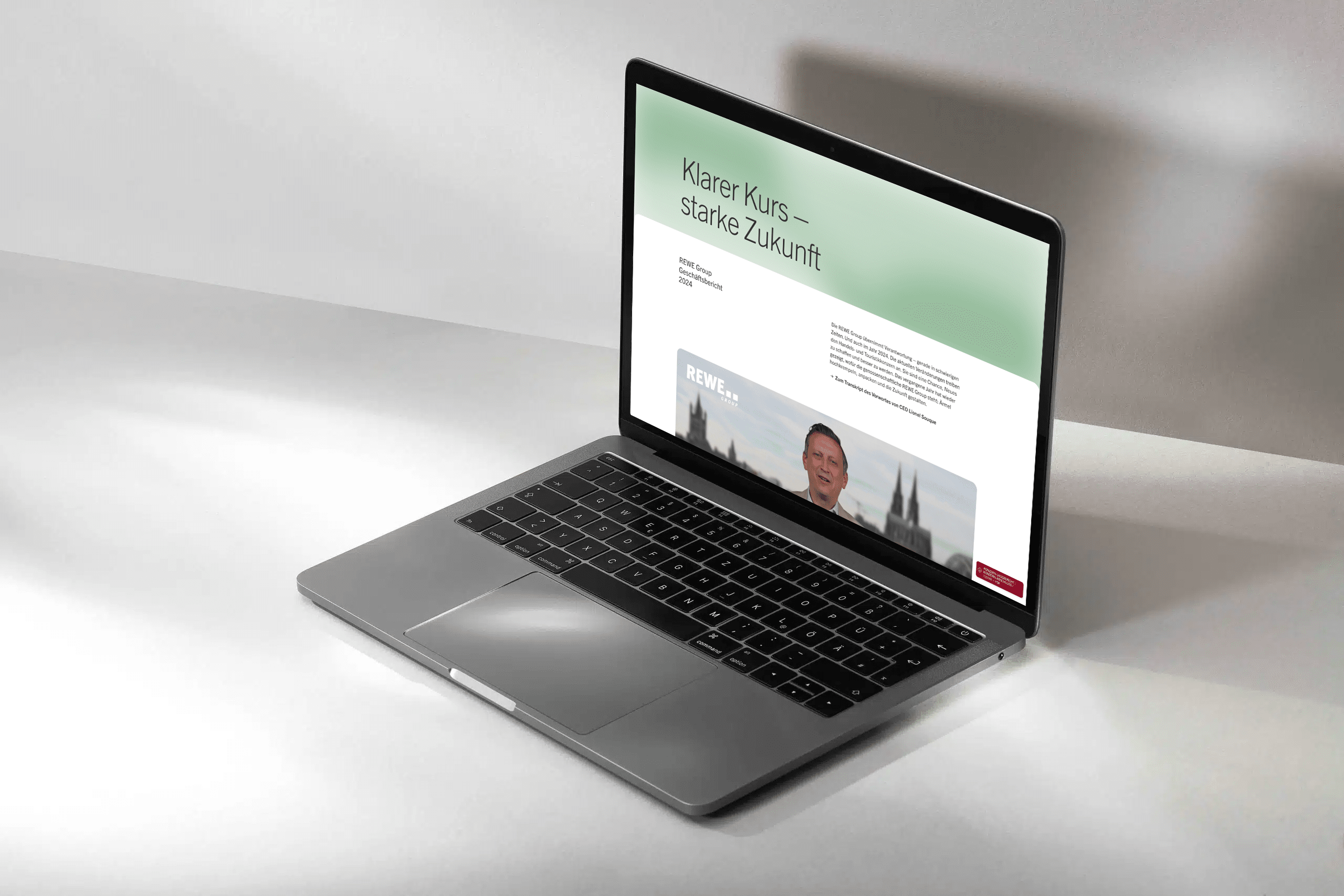 Geöffneter Laptop auf weißem Untergrund. Auf dem Bildschirm ist die Startseite des Geschäftsberichts der REWE Group 2024.
