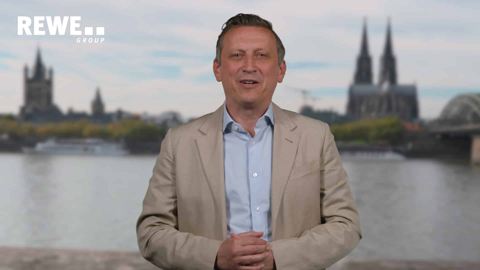 Lionel Souque, CEO der REWE Group, spricht lächelnd in die Kamera. Im Hintergrund ist das Kölner Panorama mit dem Kölner Dom und Rhein zu sehen.