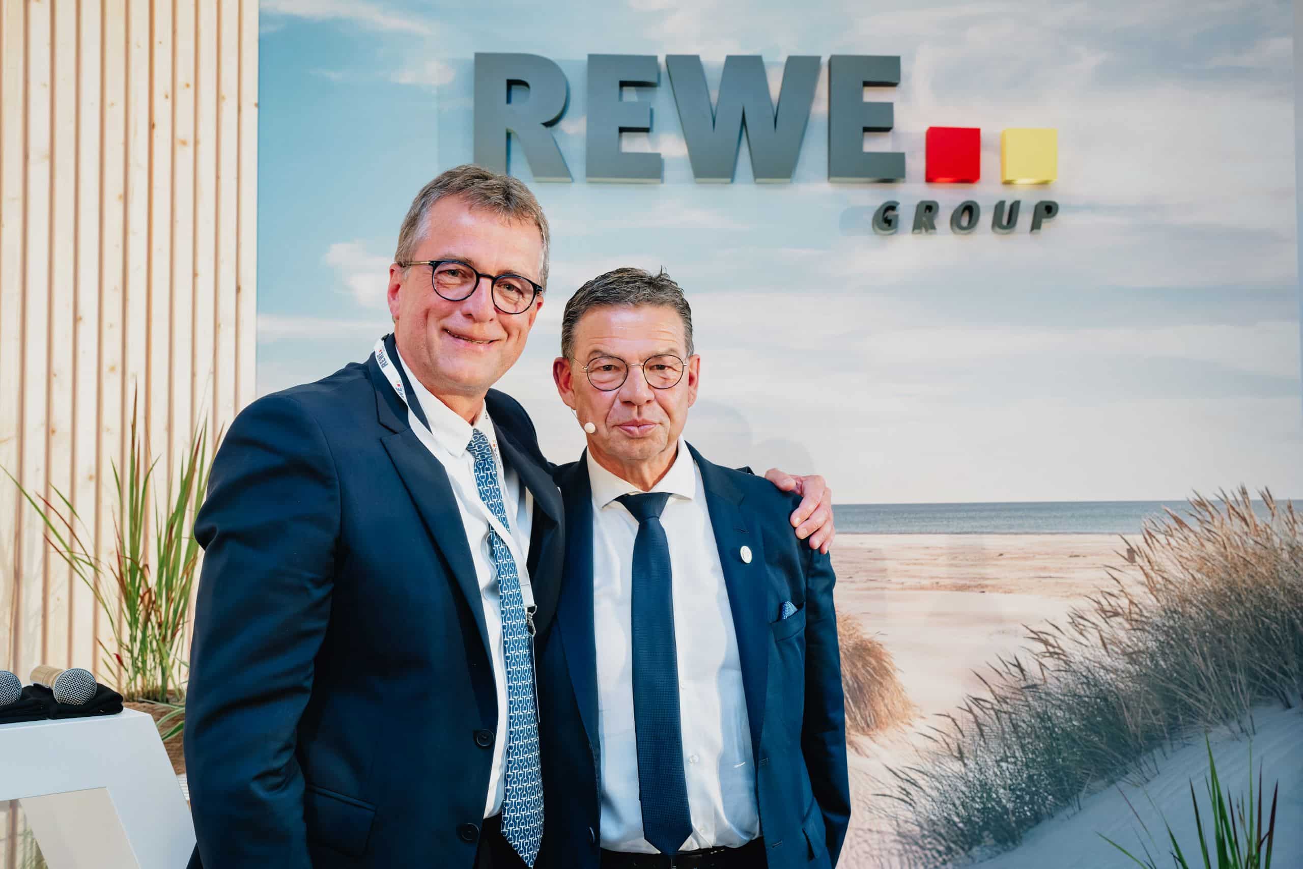 Erich Stockhausen und Stefan Lenk stehen nebeneinander und lächeln in die Kamera. Im Hintergrund ist ein Wandbild einer Düne und das REWE Group Logo.
