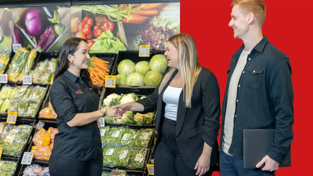 Drei Personen stehen in einem REWE Markt und lächeln sich an. Zwei Personen geben sich die Hand. Der Hintergrund ist zur Hälfte rot und zur anderen Hälfte die Gemüseabteilung des Marktes.