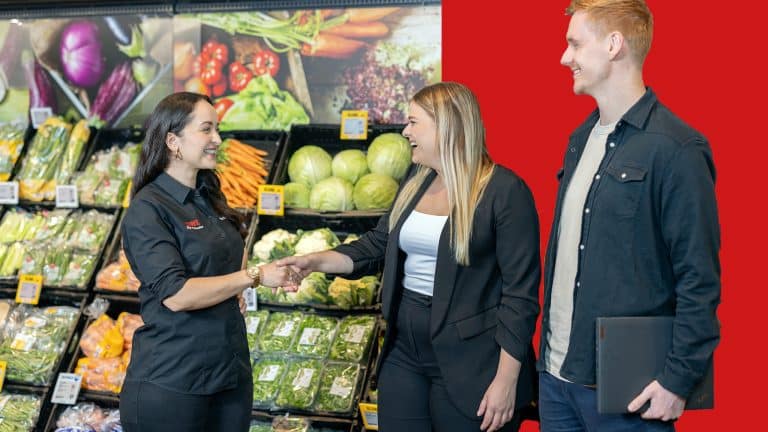 Drei Personen stehen in einem REWE Markt und lächeln sich an. Zwei Personen geben sich die Hand. Der Hintergrund ist zur Hälfte rot und zur anderen Hälfte die Gemüseabteilung des Marktes.