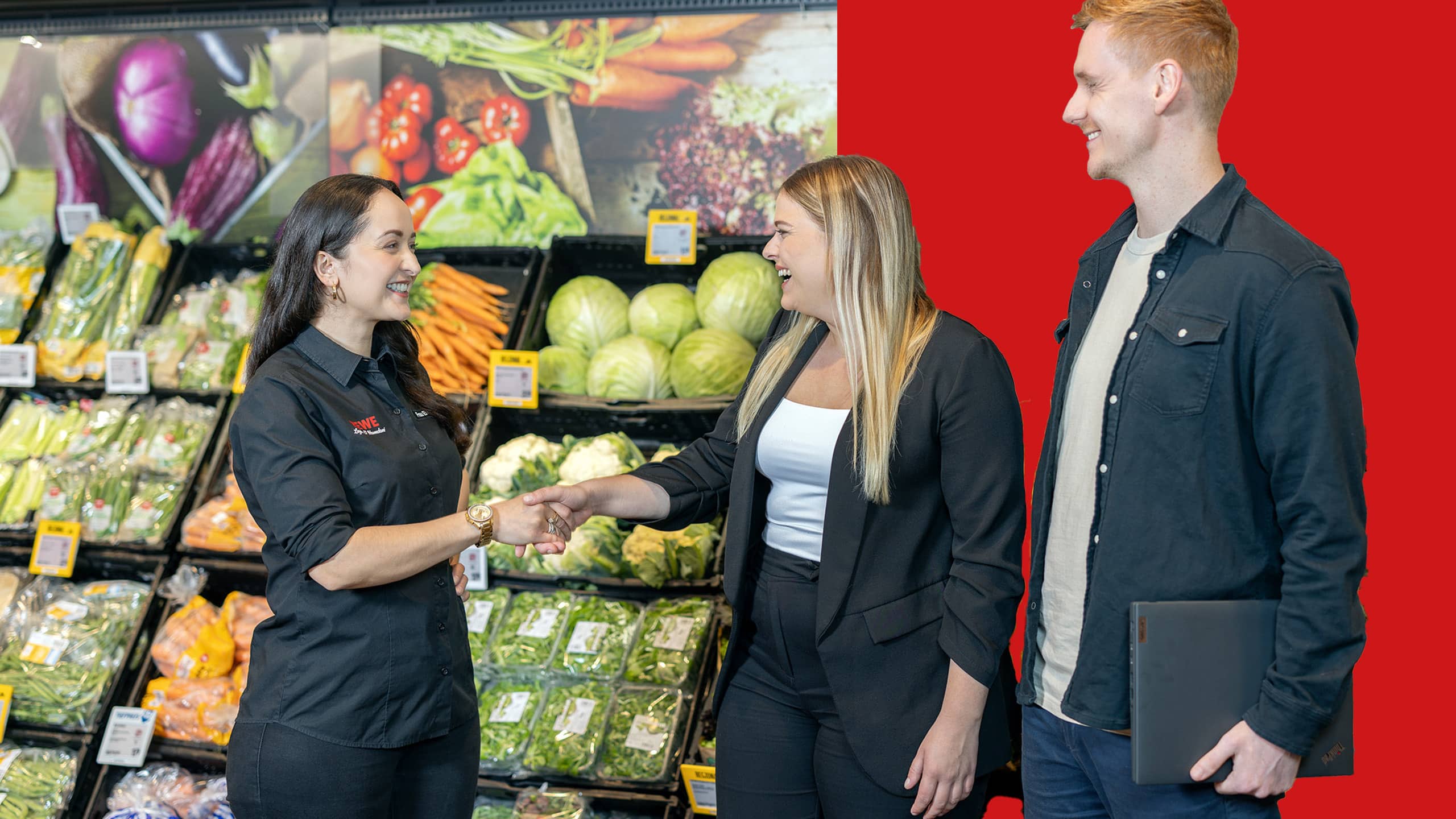 Three people are standing in a REWE supermarket and smiling at each other. Two people are shaking hands. Half of the background is red and the other half shows the supermarket's vegetable section.