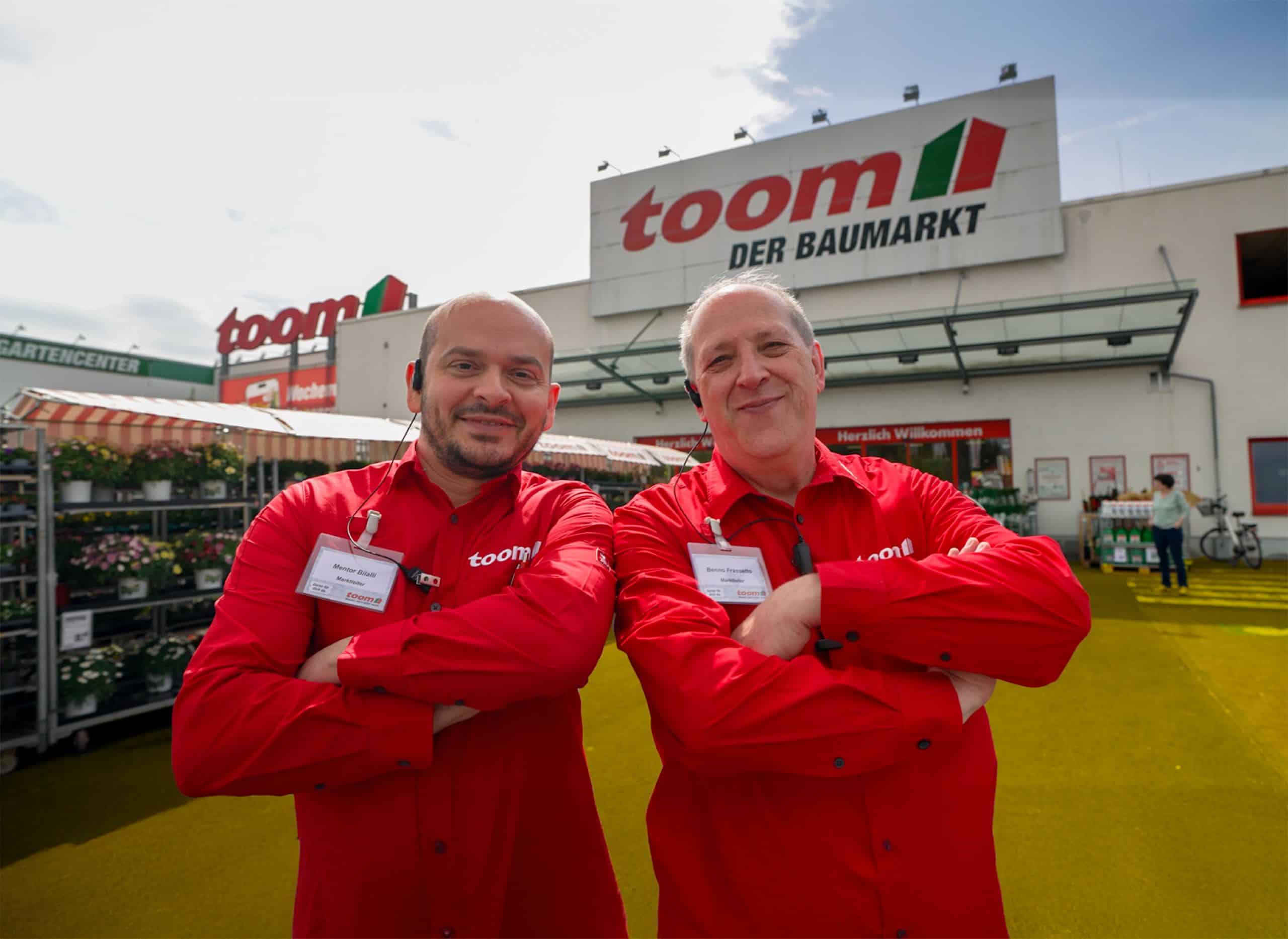 Benno Frassetto and Mentor Bilalli stand next to each other with their arms crossed and smile into the camera. A toom Baumarkt DIY store can be seen in the background from the outside.
