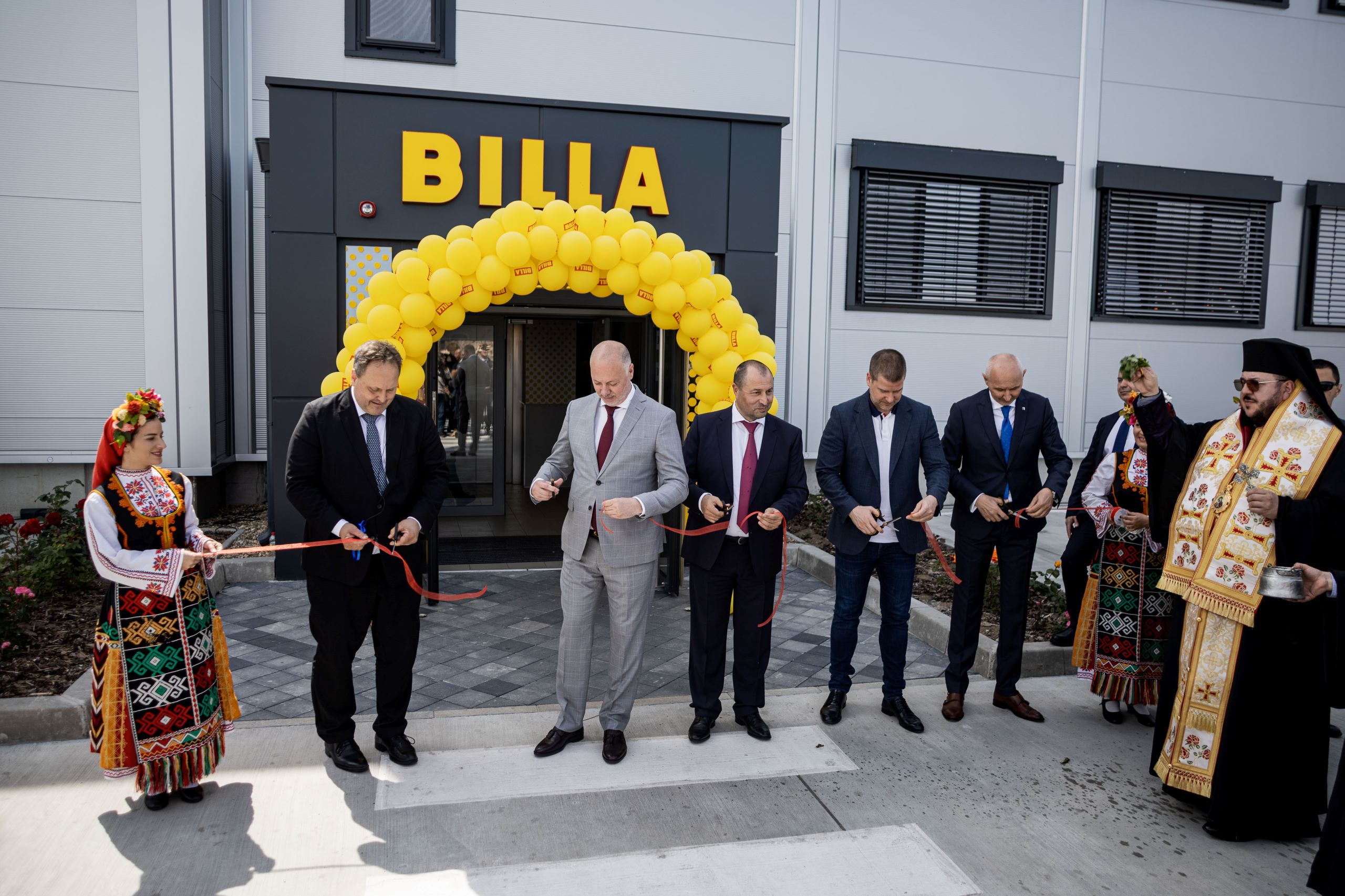 A row of people stand in front of the new BILLA CEE store in Bulgaria and cut a red ribbon to mark the opening. The entrance is decorated with yellow BILLA balloons.