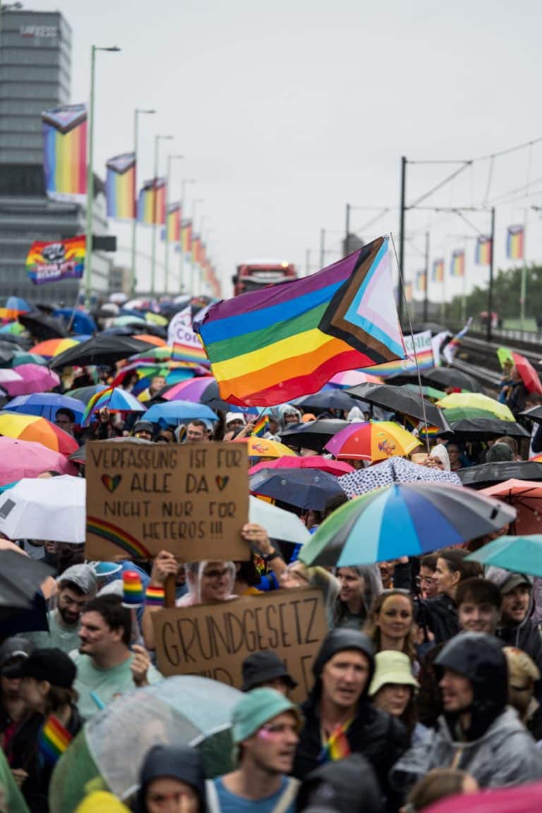 Menschenmenge beim CSD in Köln mit Regenbogenflaggen und Plakaten. Im Mittelpunkt ist eine große, wehende Pride Flagge zu sehen.