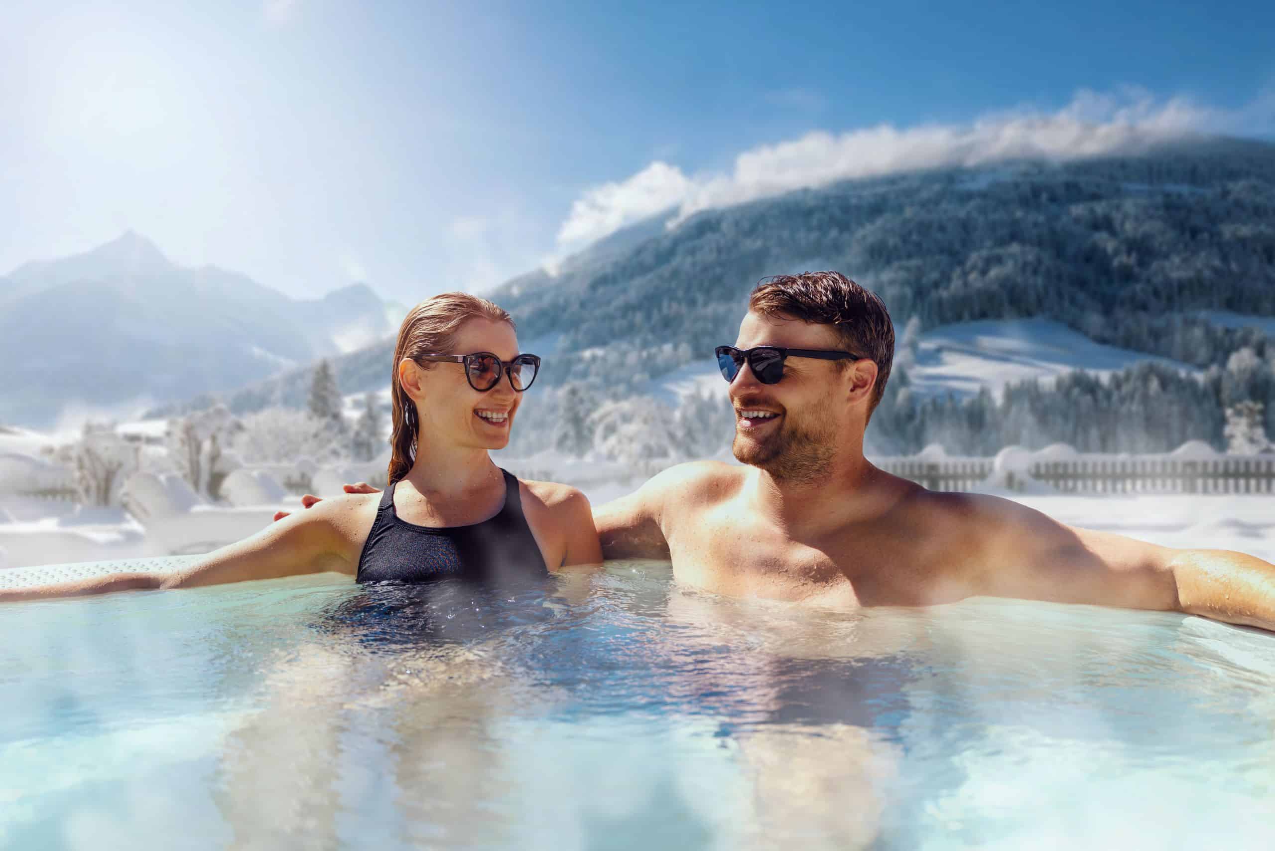 Two people wearing sunglasses are sitting in a steaming pool. In the background are snow-covered mountains.