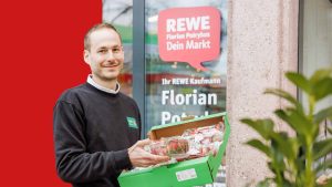 Portrait of Konstantin Tönnies. He is holding a box of strawberries and smiling at the camera. The entrance to a REWE store can be seen in the background and a red bar covers part of the background.