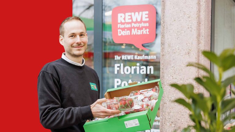 Portrait of Konstantin Tönnies. He is holding a box of strawberries and smiling at the camera. The entrance to a REWE store can be seen in the background and a red bar covers part of the background.