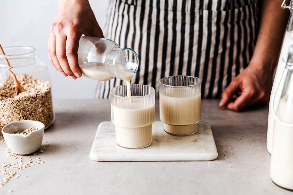 Zwei mit pflanzlichem Drink gefüllte Gläser stehen nebeneinander, in das eine Glas schenkt eine Person aus einer Karaffe den Pflanzendrink ein. Daneben steht ein Glasbehälter mit Haferflocken.