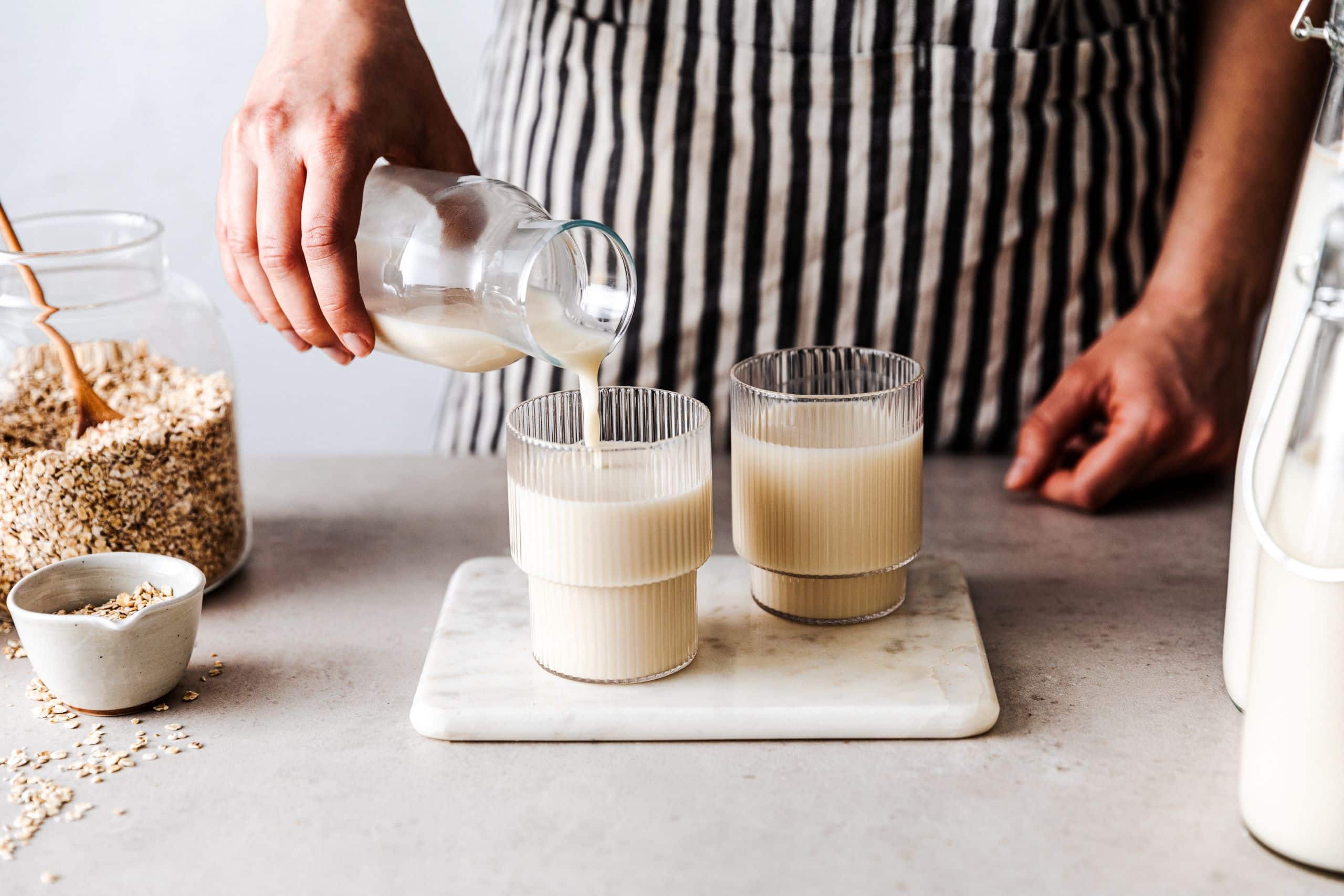 Zwei mit pflanzlichem Drink gefüllte Gläser stehen nebeneinander, in das eine Glas schenkt eine Person aus einer Karaffe den Pflanzendrink ein. Daneben steht ein Glasbehälter mit Haferflocken.