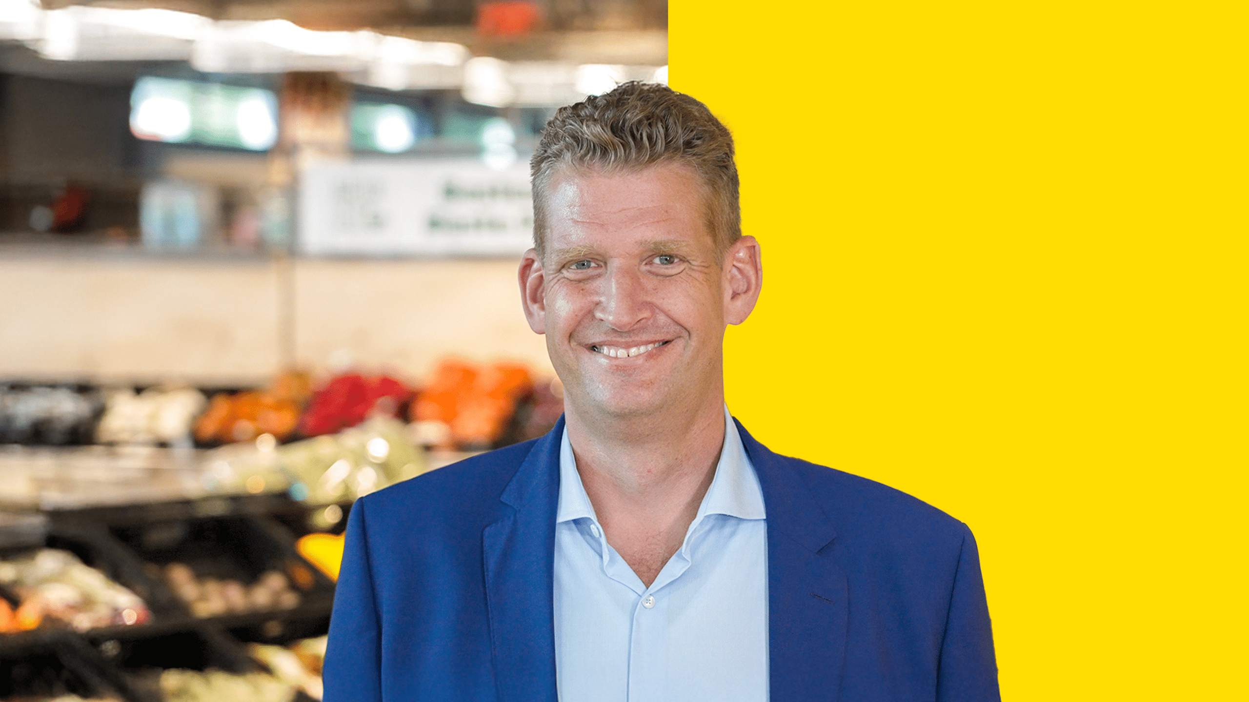 Portrait of Stefan Görgens. The vegetable section of a PENNY supermarket can be seen blurred in the background and a yellow bar across half of the background.
