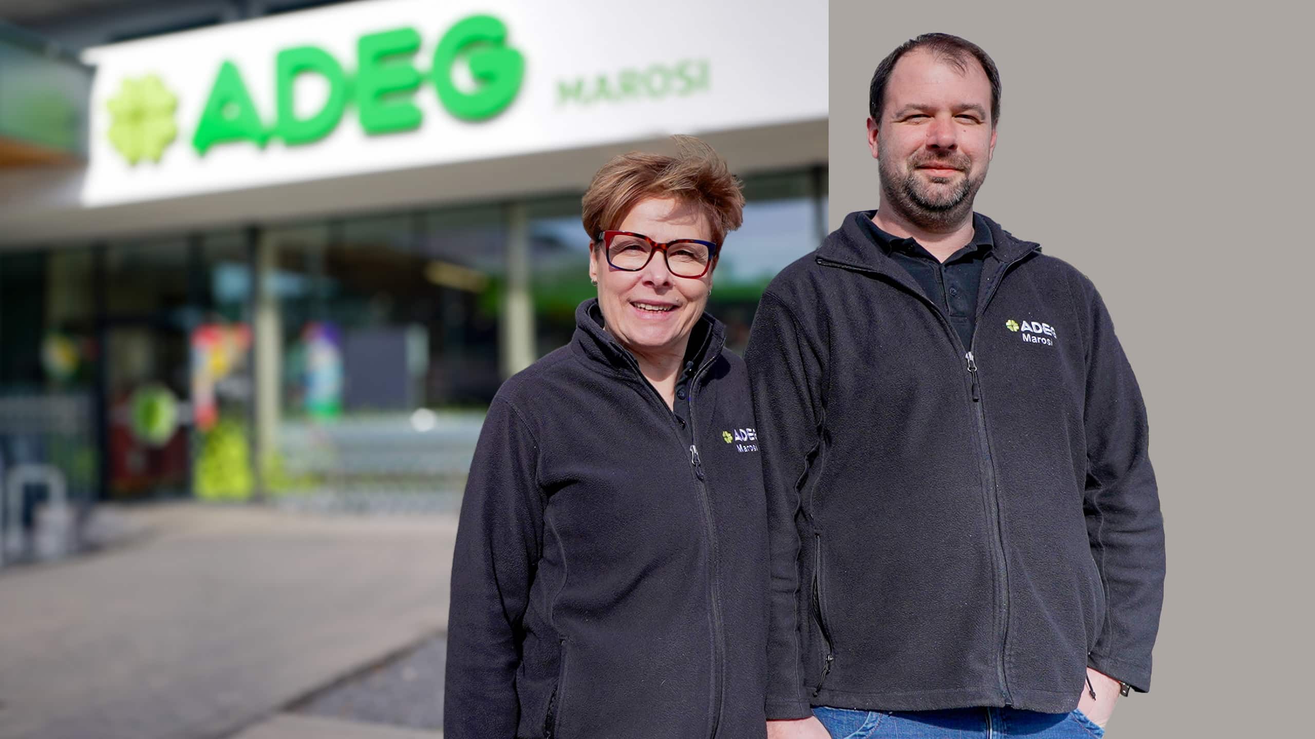 Porträt von Erna & Daniel Marosi vor ihrem ADEG Markt. Der Hintergrund ist zur Hälfte grau hinterlegt.