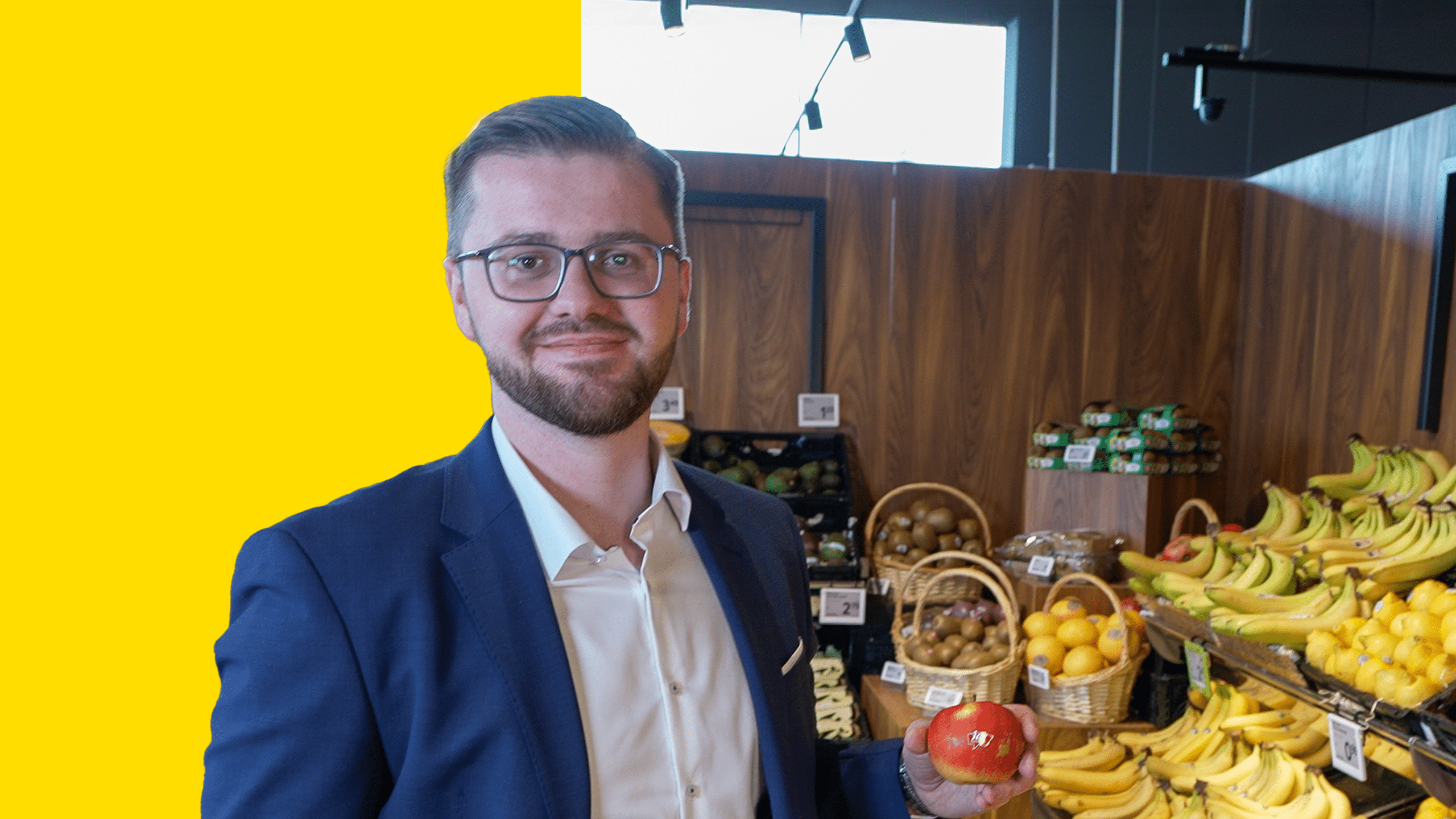 Porträt von Kurt Akalinmit einem Apfel in der Hand in der Obstabteilung eines BILLA Marktes. Der Hintergrund ist zur Hälfte mit einem gelben Balken hinterlegt.
