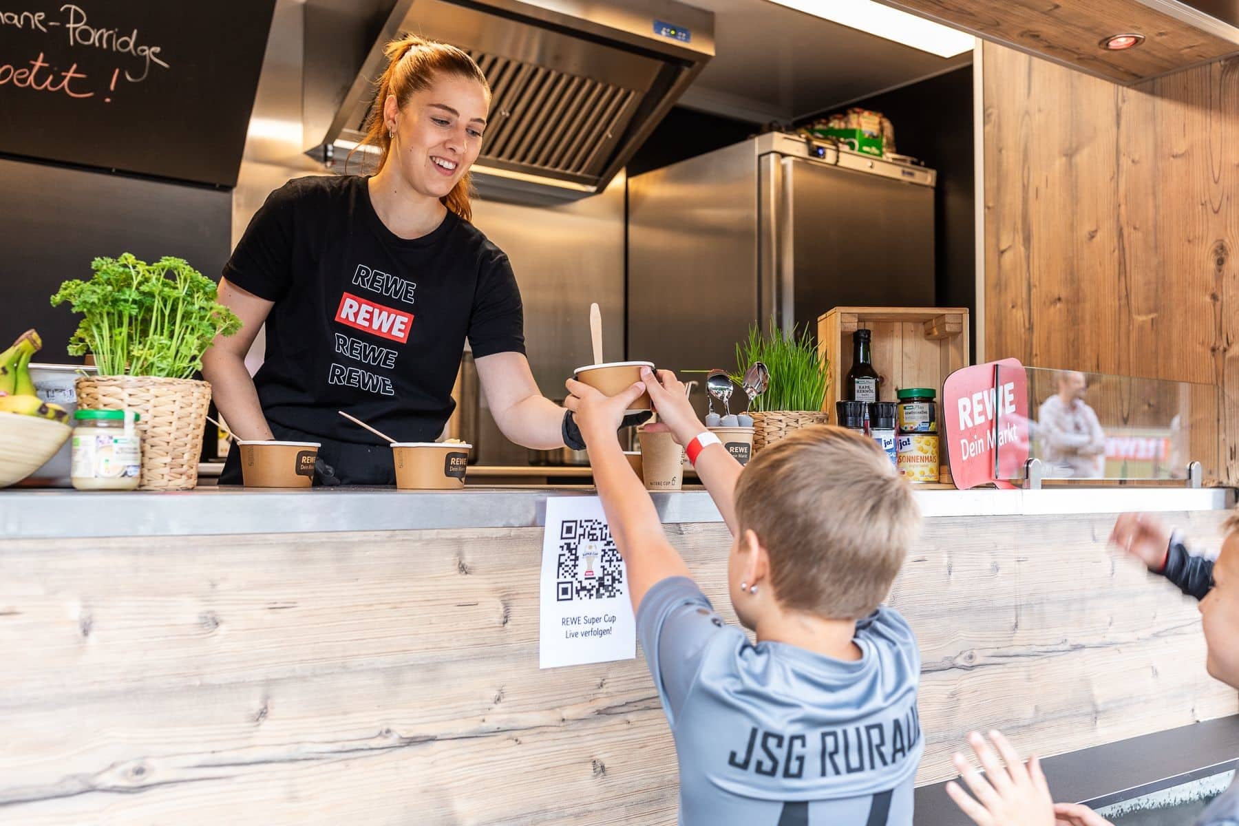 Frau die Kind Essen aus einem Foodtruck anreicht im Rahmen der REWE „Mission Foodtruck“.