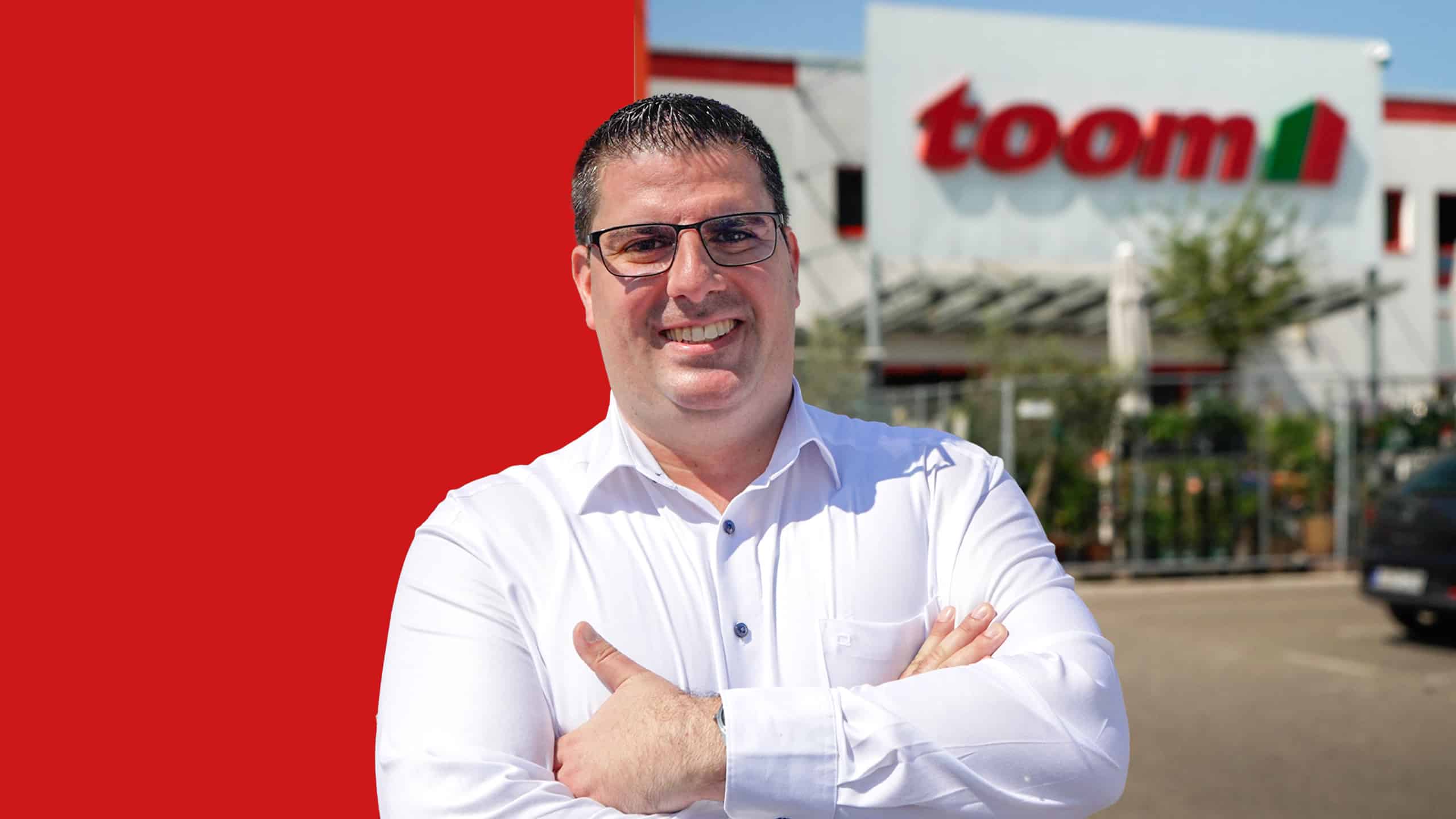 Porträt von Theodoros Lazaridis vor seinem toom Markt. Der Hintergrund ist zur Hälfte rot hinterlegt.