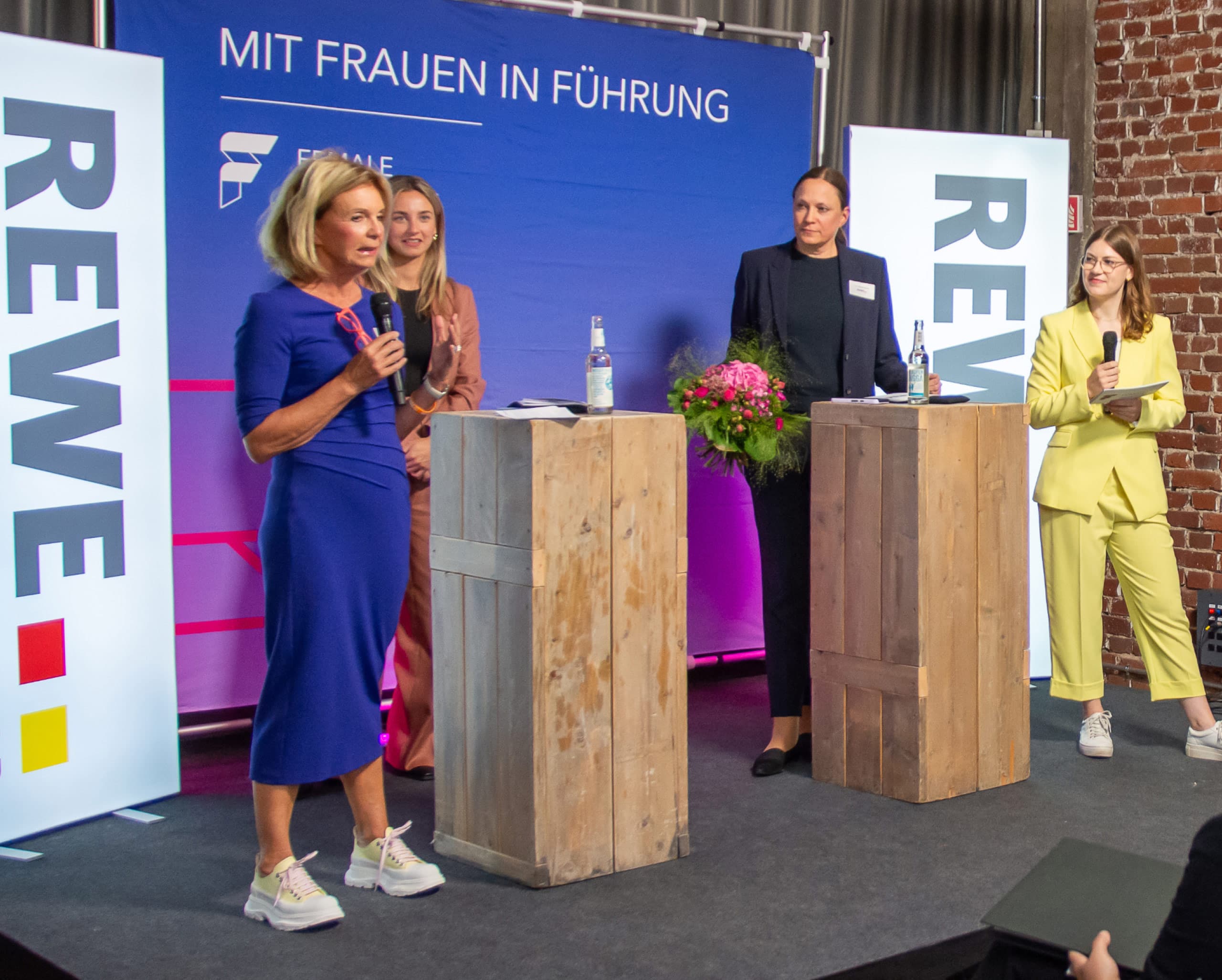 Four women are standing at a wooden desk on a stage at an event with the theme 'With women in leadership', banners with REWE logos can be seen at the edge of the stage. The women are wearing trouser suits.