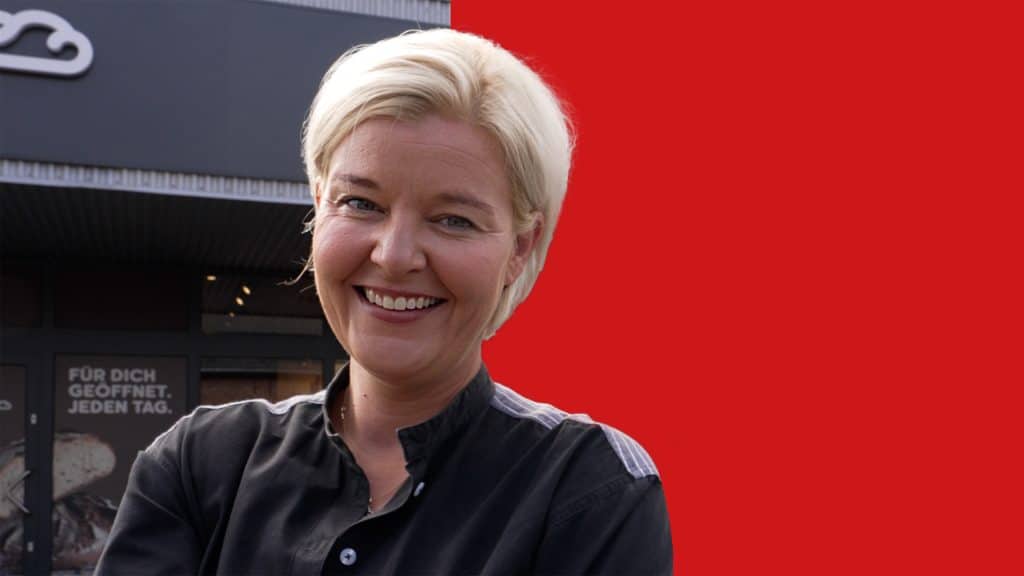 Porträt von Kathrin Goedecke vor ihrem Supermarkt. Der Hintergrund ist zur Hälfte rot hinterlegt.