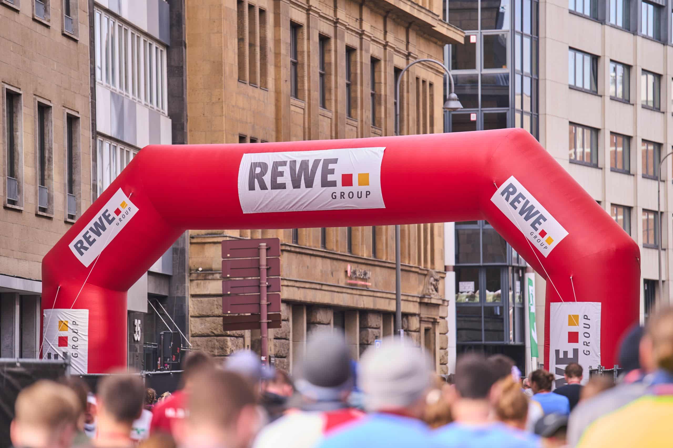 Ein aufblasbares Tor mit dem Logo der REWE Group markiert das Ziel des Köln Marathons. Zahlreiche Menschen sind um das Tor herum versammelt.