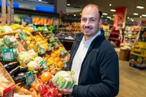 Porträt von Leon Lindemann. Er steht in der Gemüseabteilung eines REWE Marktes und hält Blumenkohl in der Hand.