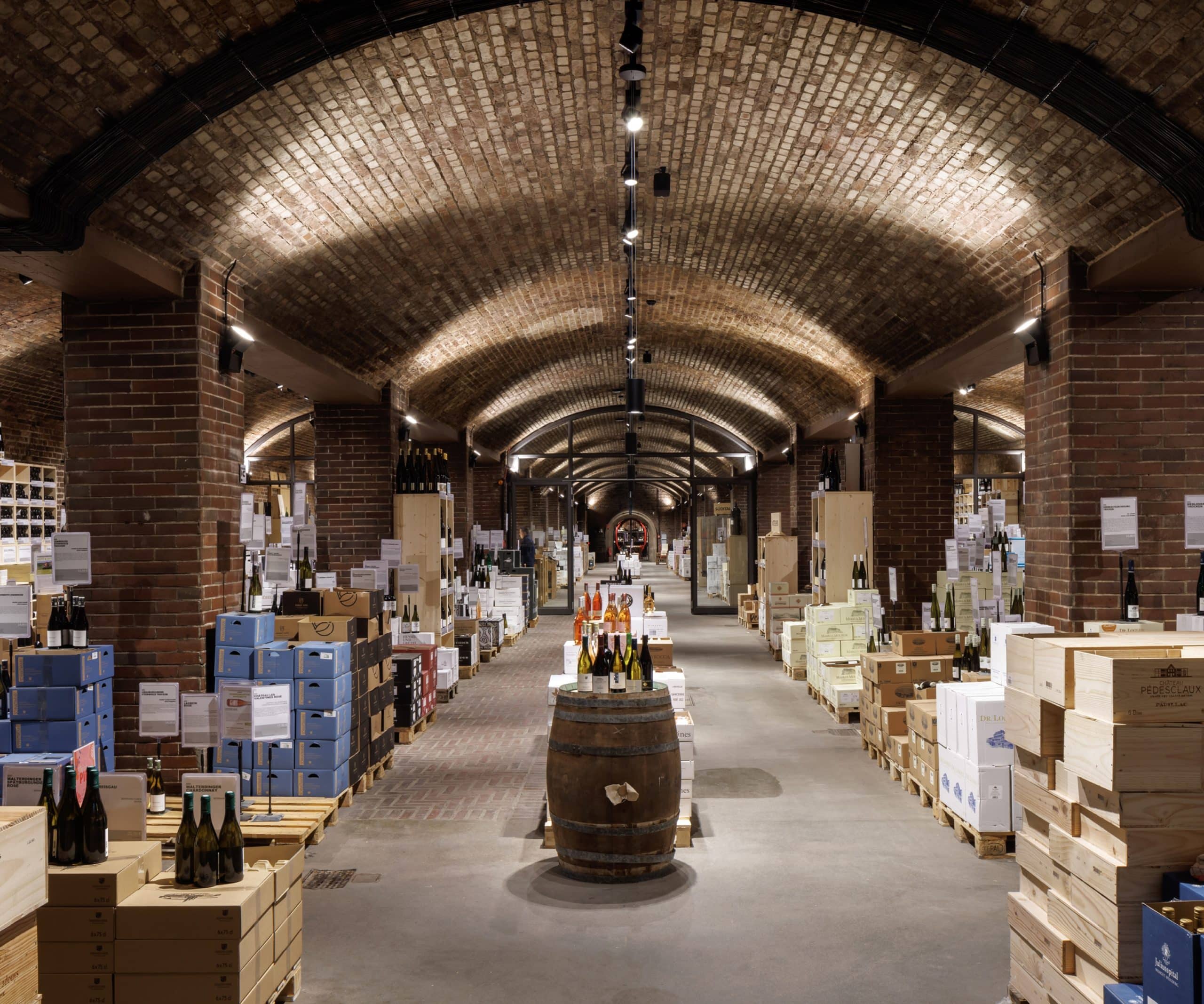 Vaulted cellar with brick arches, a wine barrel with wine bottles in the middle, stacks of wooden crates and boxes to the left and right.