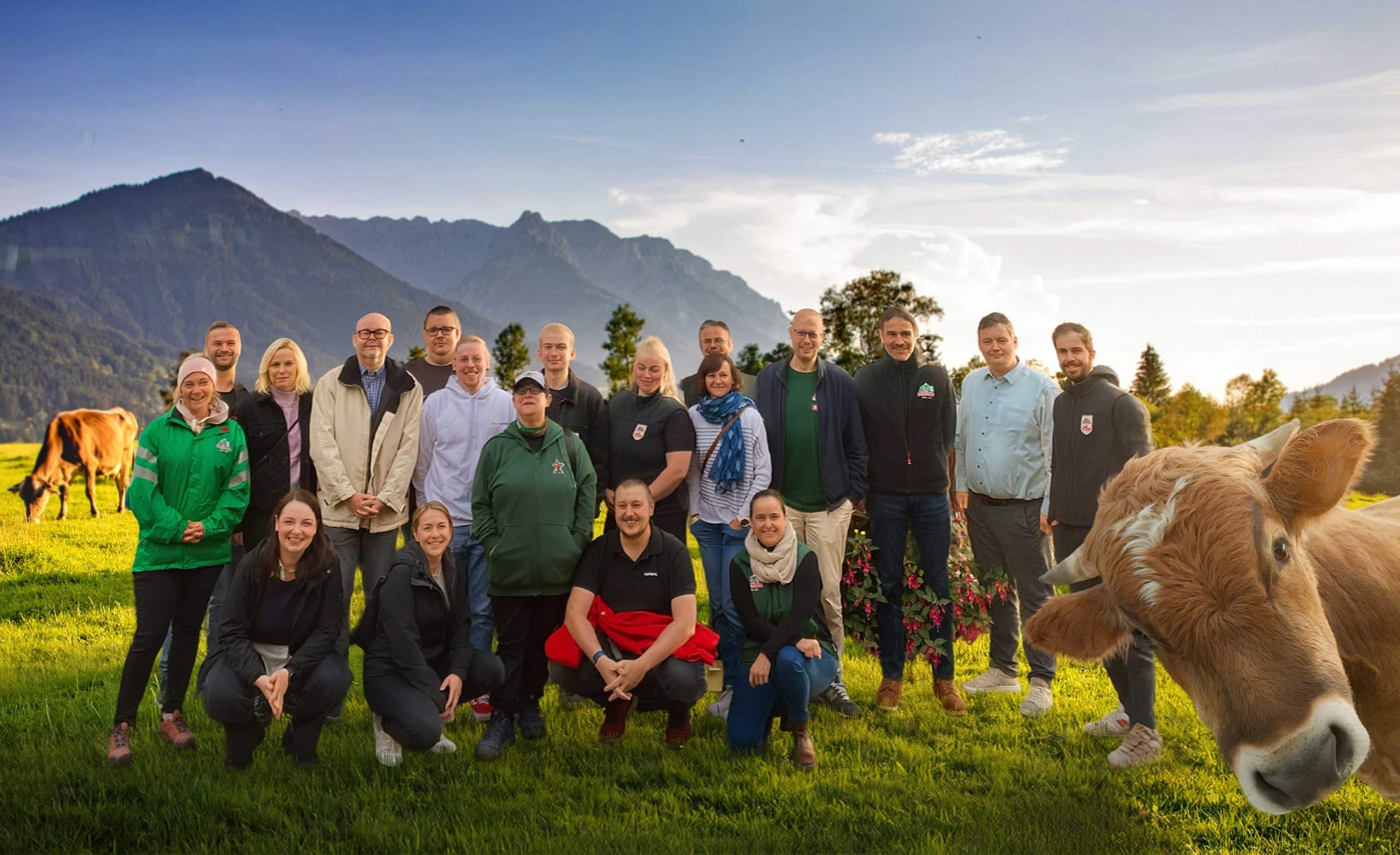 Group of people standing in a meadow with mountains in the background, to the side, at the bottom right of the picture, a brown cow looks curiously ahead.