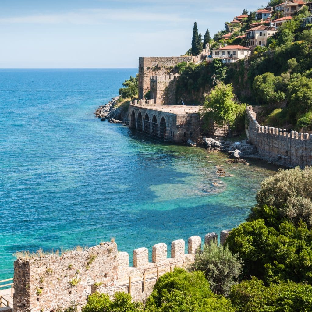 A stretch of coast with turquoise blue sea, historic stone walls and houses on a wooded slope under a blue sky.