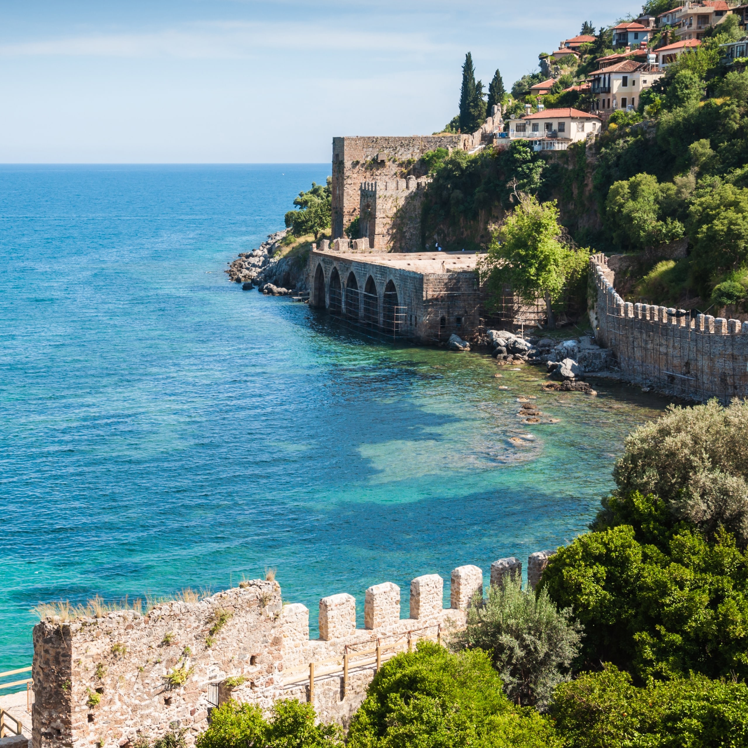 Küstenabschnitt mit türkisblauem Meer, historischen Steinmauern und Häusern auf bewaldetem Hang unter blauem Himmel.