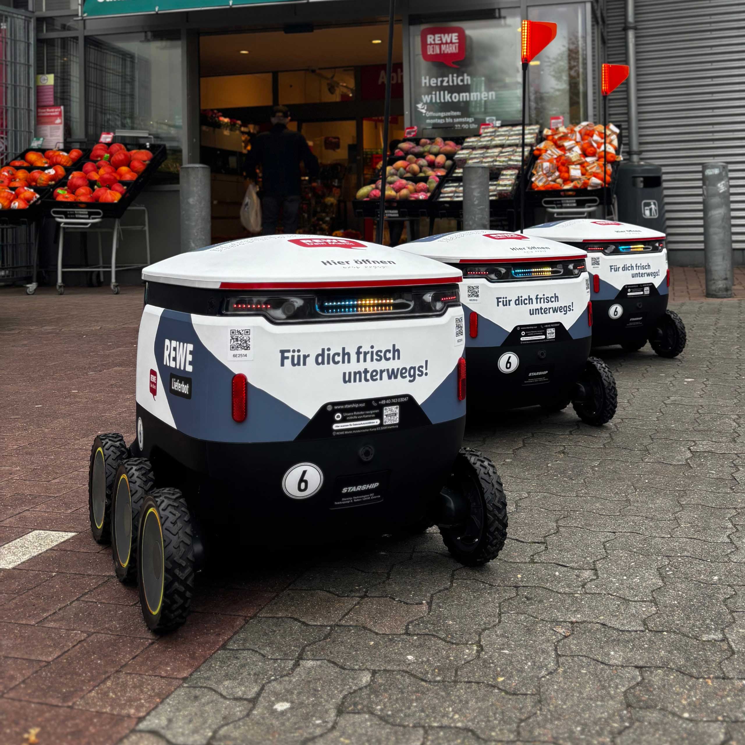 Drei Lieferroboter mit der Aufschrift Für dich frisch unterwegs! Und dem REWE Logo, stehen vor einem offenem Supermarkt.