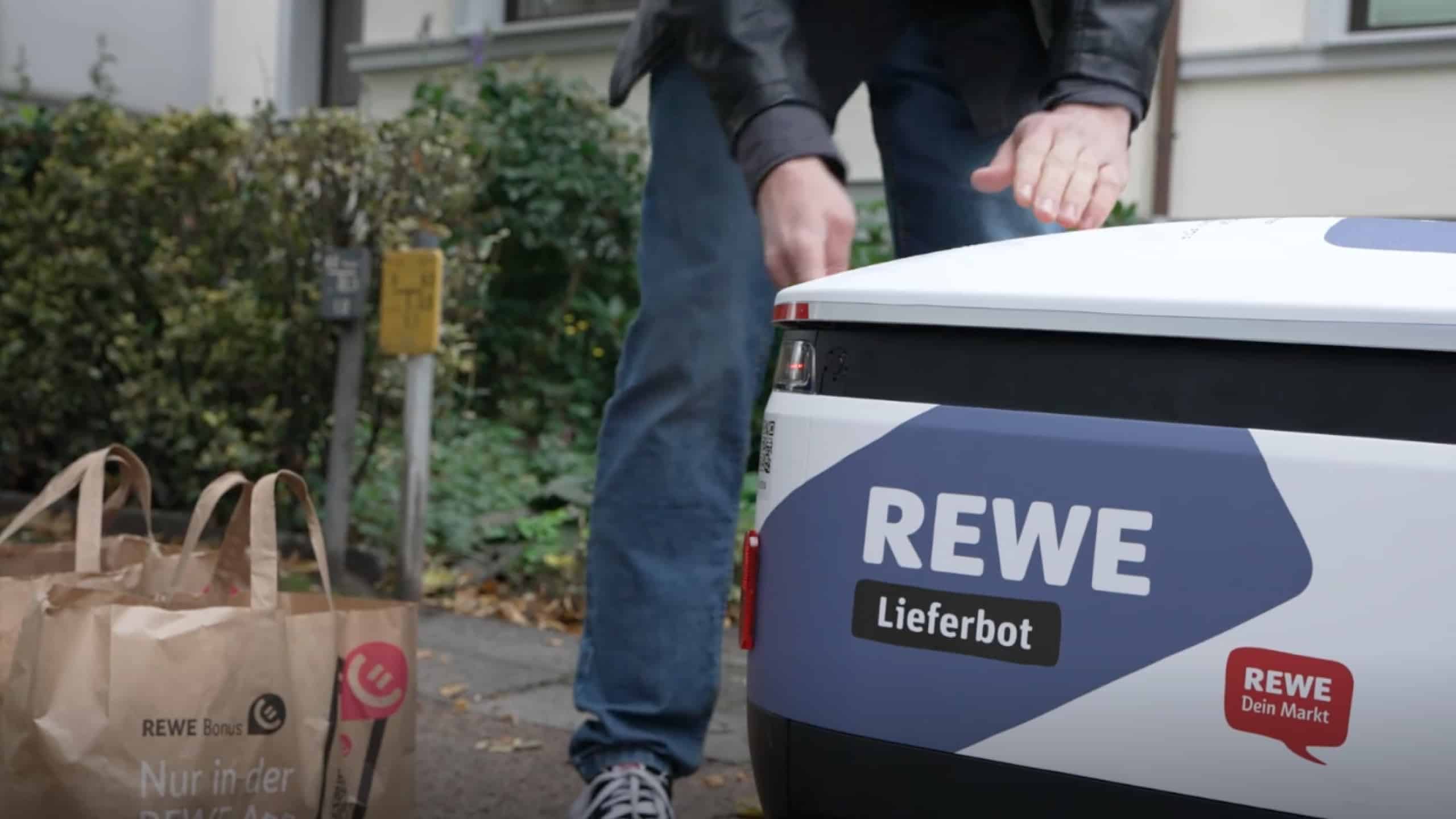 Ein Lieferroboter mit der Aufschrift REWE, der Einkaufstüten bringt.