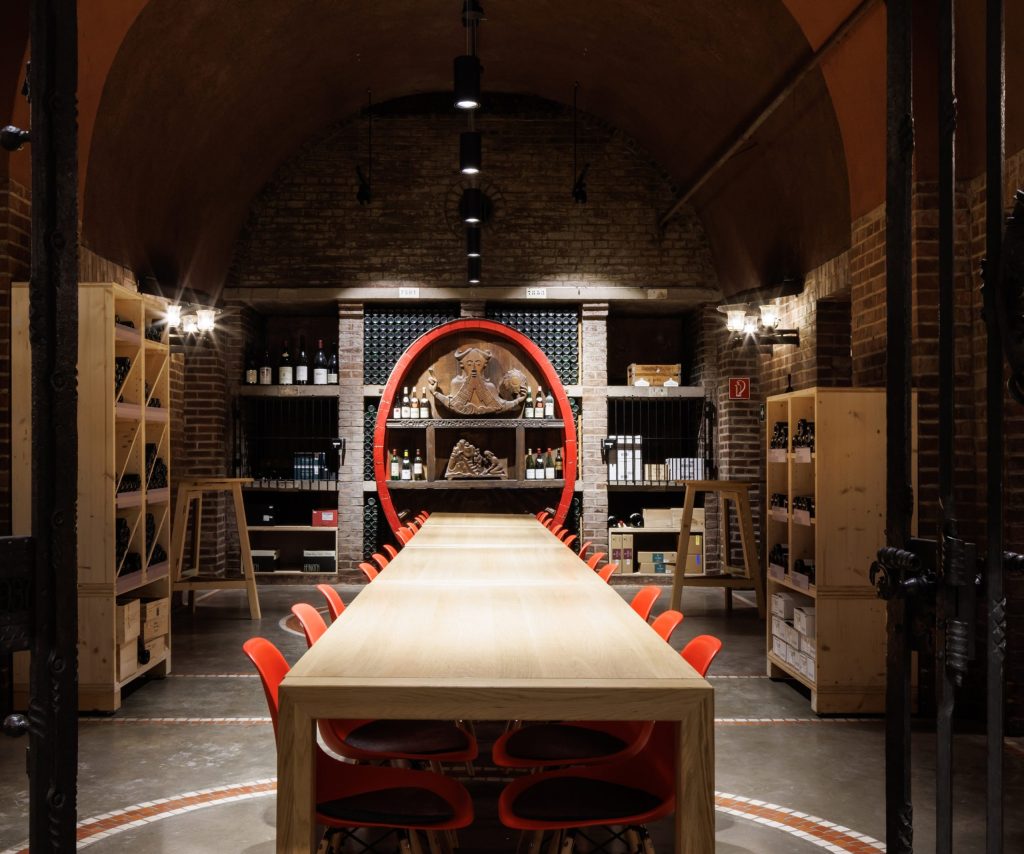 Long wooden table with red chairs in a vaulted cellar, surrounded by shelves with wine bottles and wooden paneling.