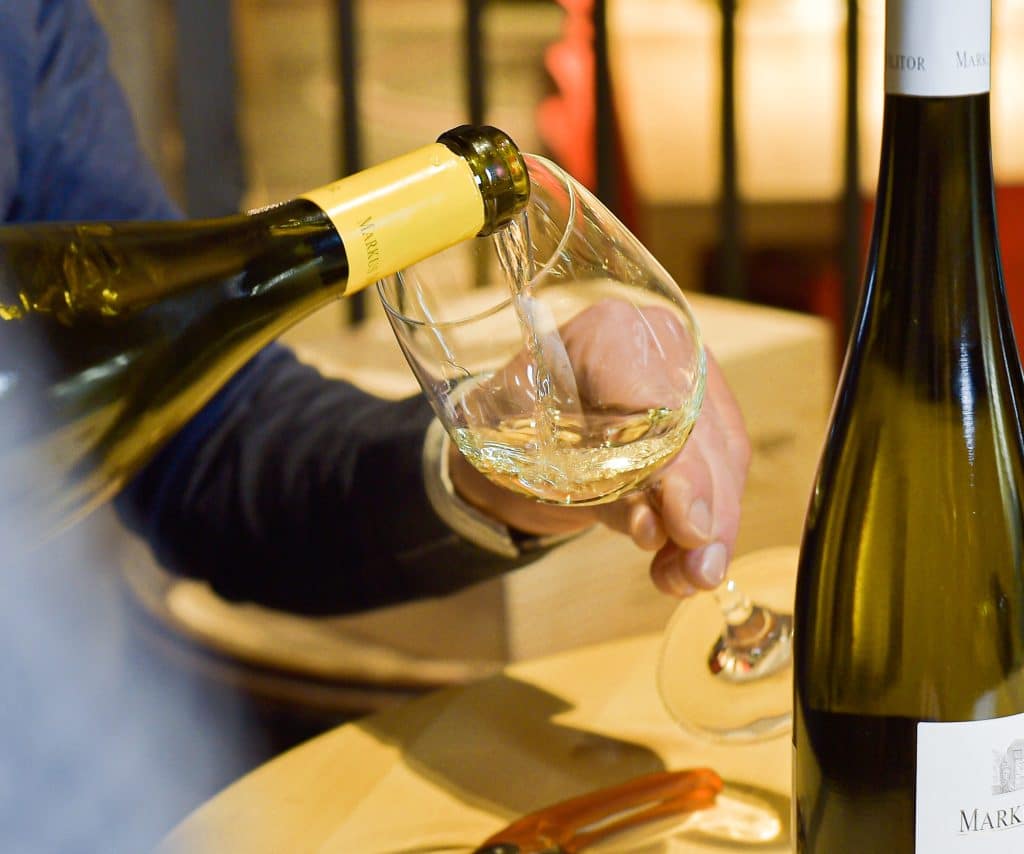 Weißwein wird aus einer Flasche mit gelbem Verschluss in ein Weinglas eingeschenkt, weitere Weinflasche steht auf dem Tisch.