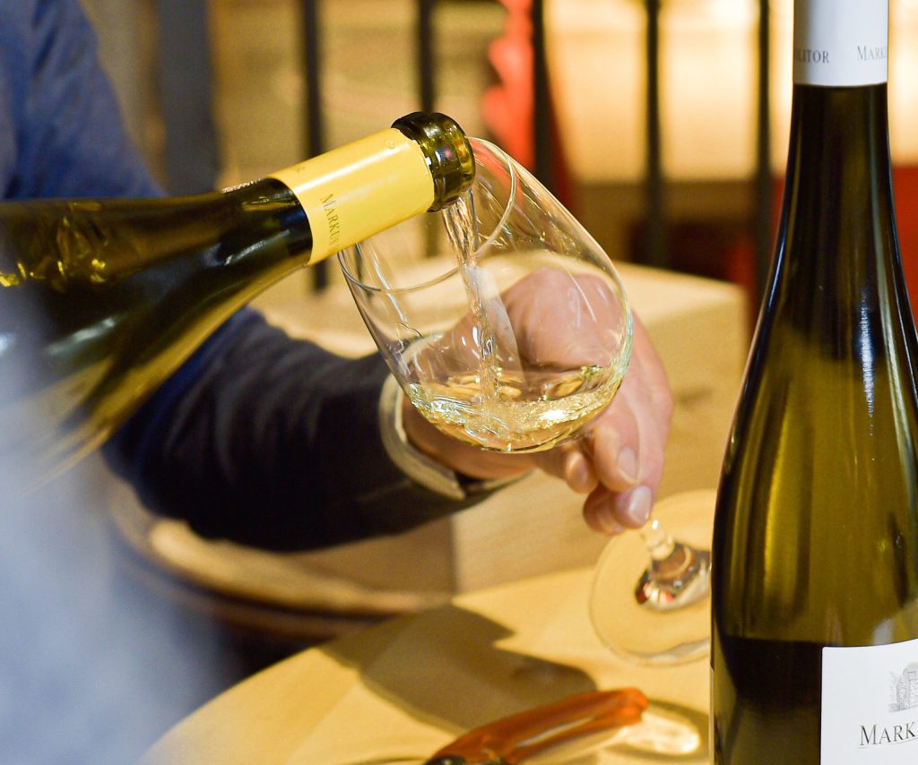 White wine is poured from a bottle with a yellow cap into a wine glass, another bottle of wine is on the table.