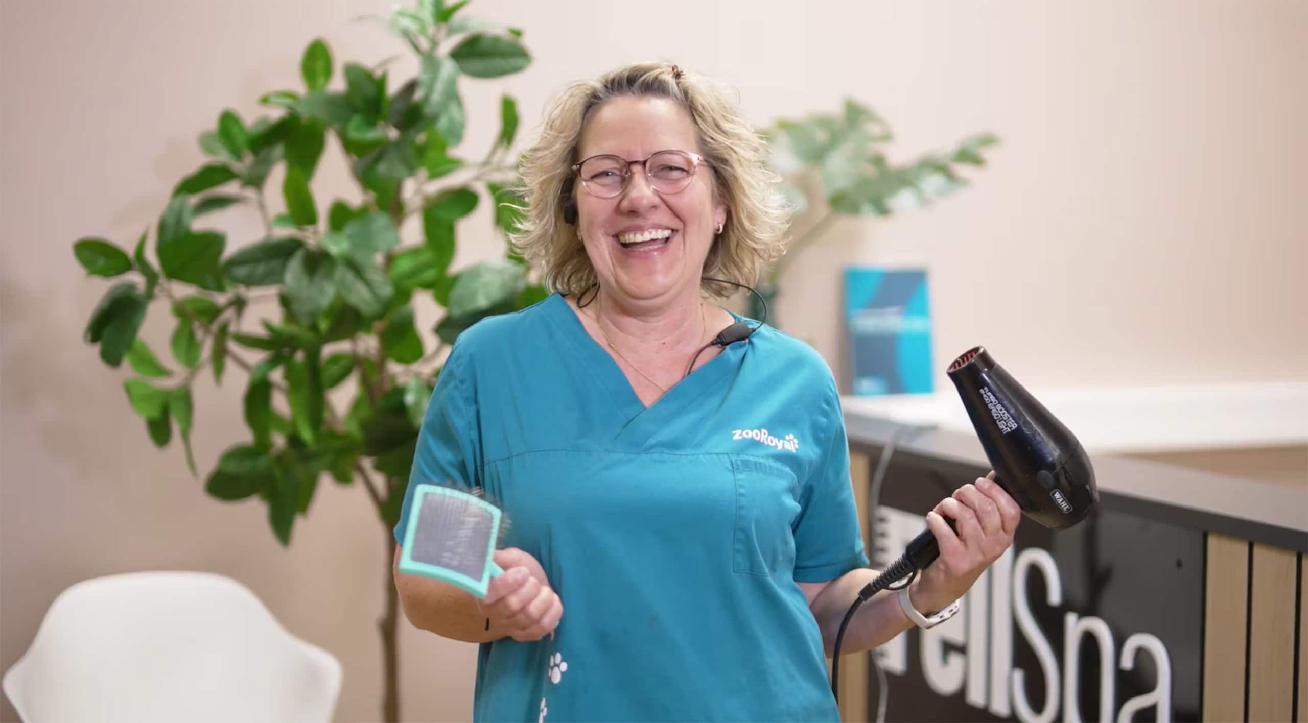 Andrea Ozekker poses with a hair dryer and comb in a blue smock with the words “ZooRoyal” written on it.