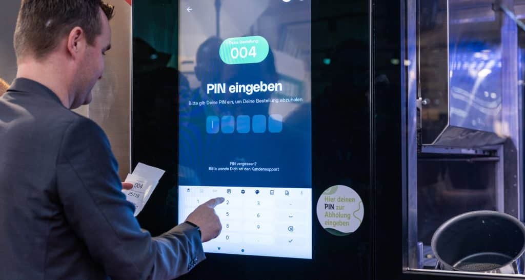 Mann in dunklem Anzug gibt PIN auf großem Touchscreen ein, daneben Roboterarm in Glasgehäuse sichtbar.