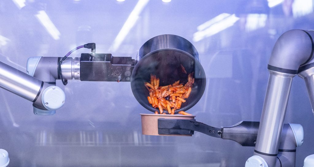 Two silver robotic arms, one tilts a pot of cooked pasta into a bowl, the other holds the bowl.