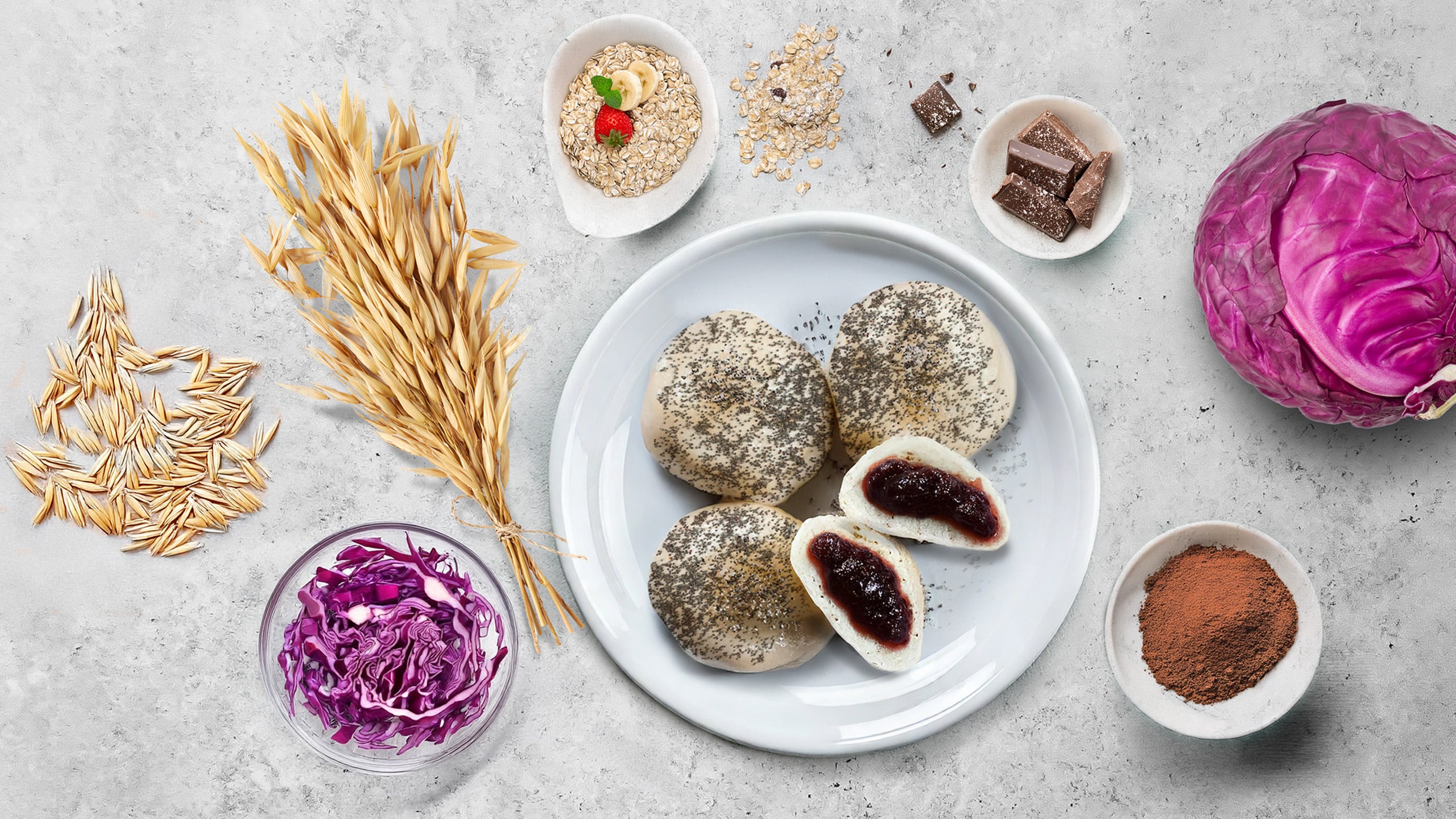 Four round pieces of dough sprinkled with poppy seeds, one cut in half and filled with red fruit filling on a white plate, next to it oats, chocolate, and red cabbage in a bowl.