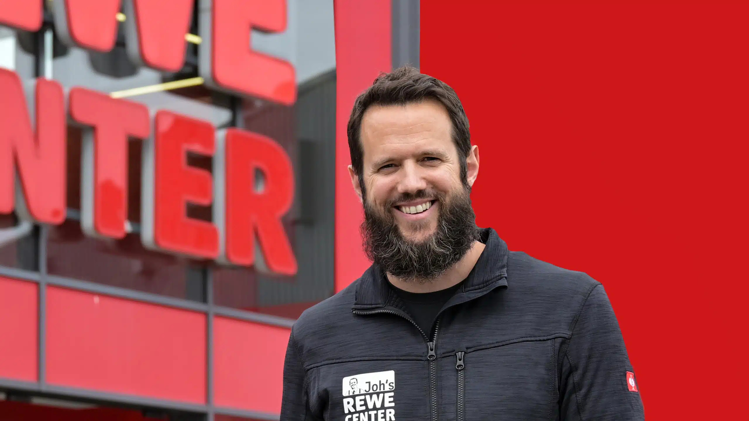 Johannes Fieber, with a dark beard, wears a black jacket with the words “Joh's REWE CENTER” in front of a red building with large letters.