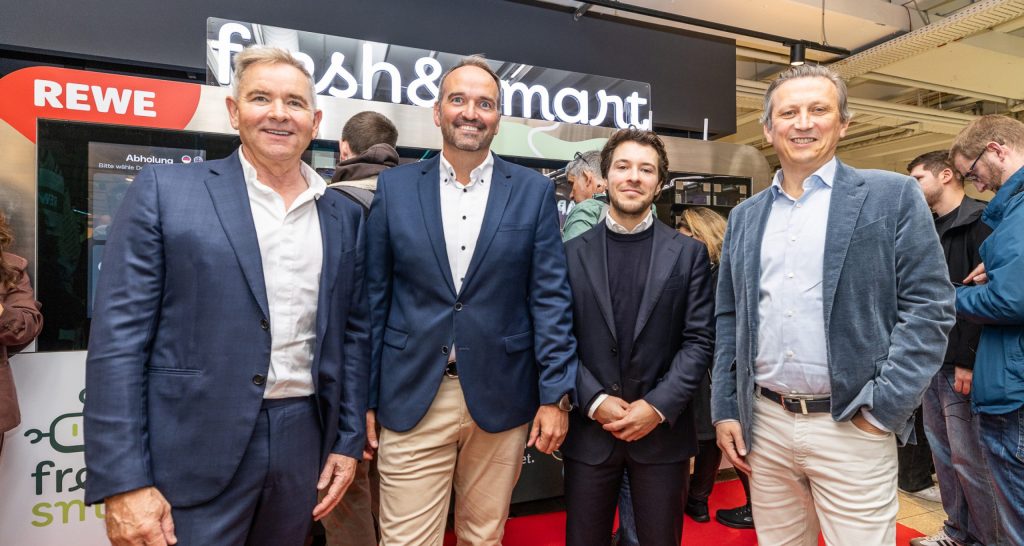 Four men in suits and shirts stand in front of a stand with the words 'fresh&smart' and a red REWE logo.