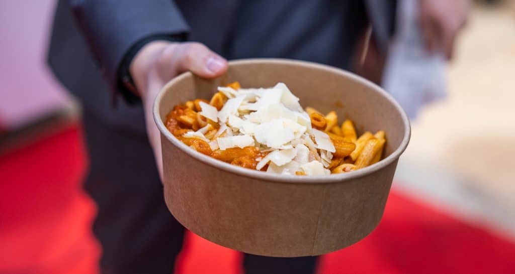 Braune Papierschale mit Penne in Tomatensauce, darüber gehobelte Käsescheiben, gehalten von einer Person in dunklem Anzug.
