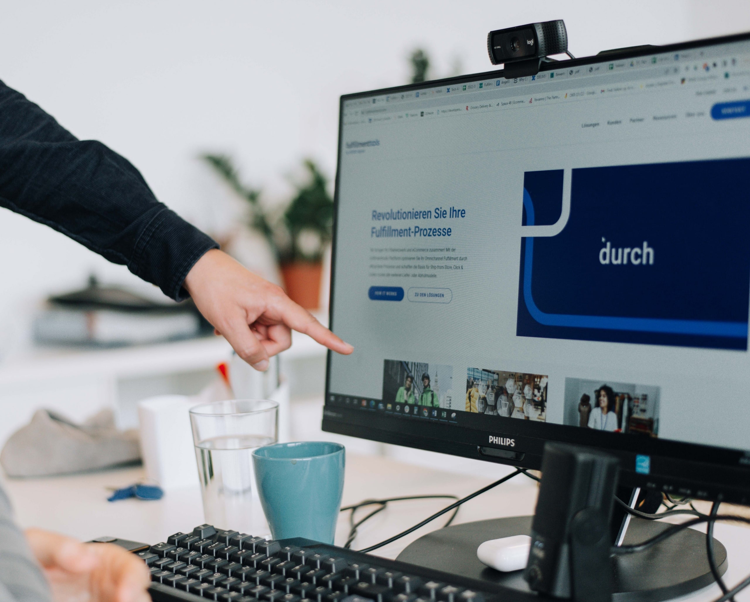 Eine Situation im Büro, wo eine Hand auf einen Monitor zeigt, der eine Website abbildet, auf der ein Text zu sehen ist: 'Revolutionieren Sie Ihre Fulfillment-Prozesse'.