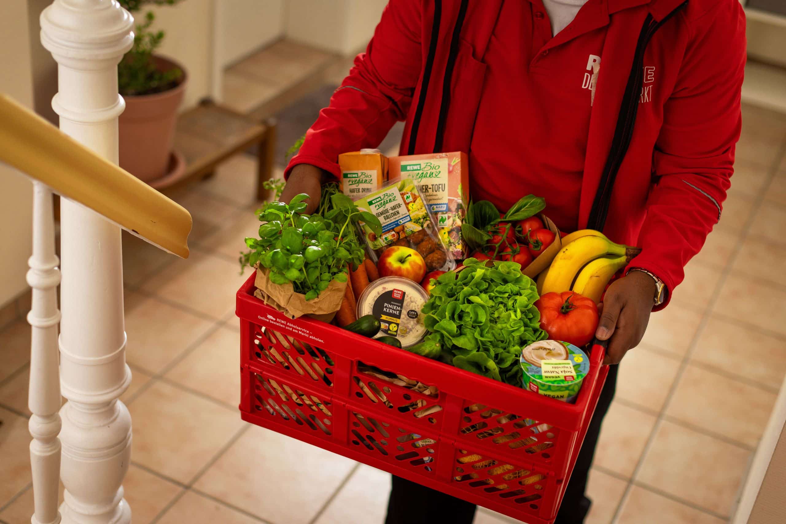 Eine Person in roter Arbeitskleidung trägt eine rote Kiste voller frischer Lebensmittel wie Obst, Gemüse, Salat und Kräuter in einem Treppenhaus.