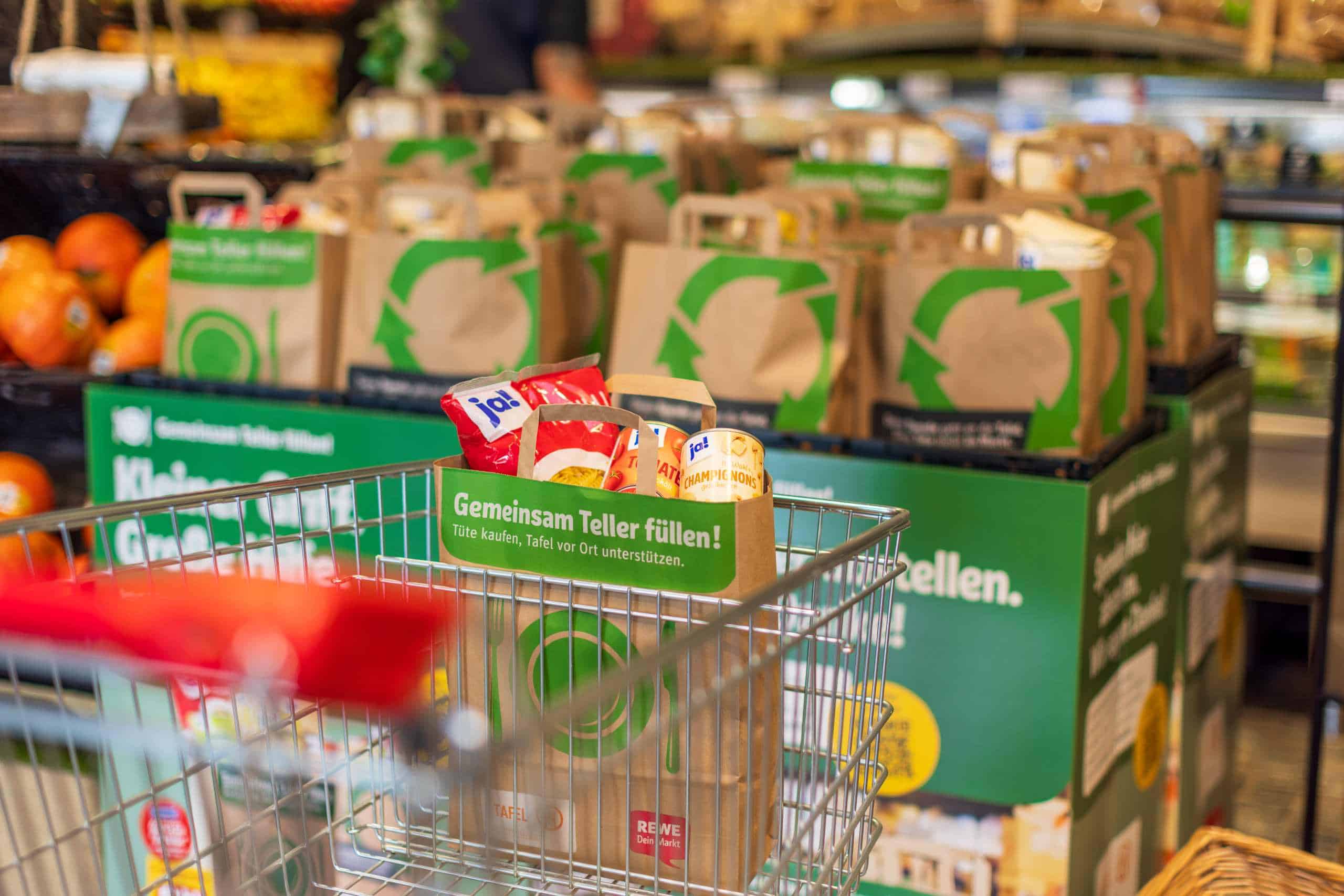 A shopping cart with long-life foodstuffs such as pasta and flour stands in the foreground of a supermarket. Behind it, many brown paper bags with recycling symbols are piled up. There is a green sign on the shopping cart advertising a donation campaign for the food bank.