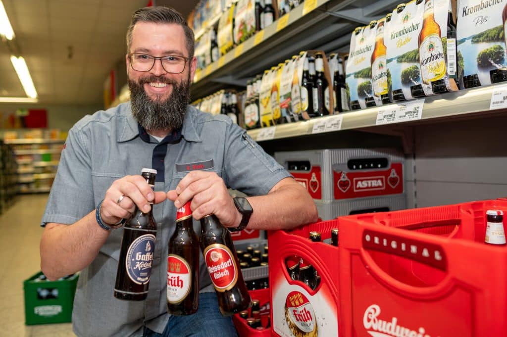 Der REWE Kaufmann, Sebastian Stein, steht vor Regalen mit Bier und hält in die Händen drei Flaschen von verschiedene Biersorten.